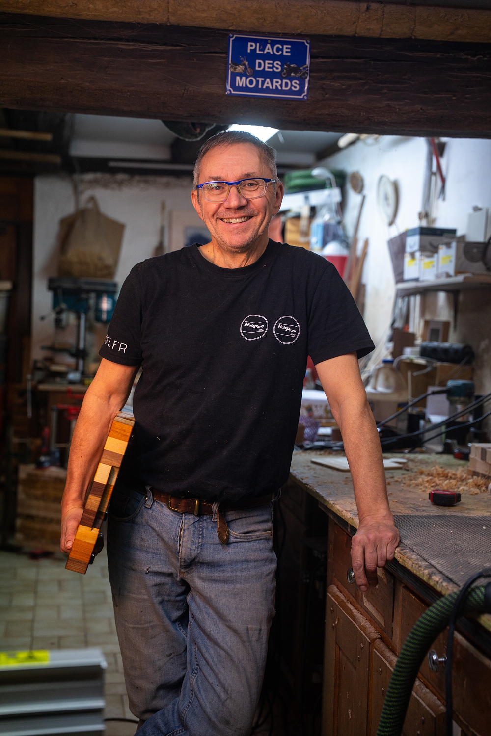Homme souriant portant des lunettes bleues et un t-shirt noir dans un atelier de menuiserie, tenant une planche en bois multicolore.