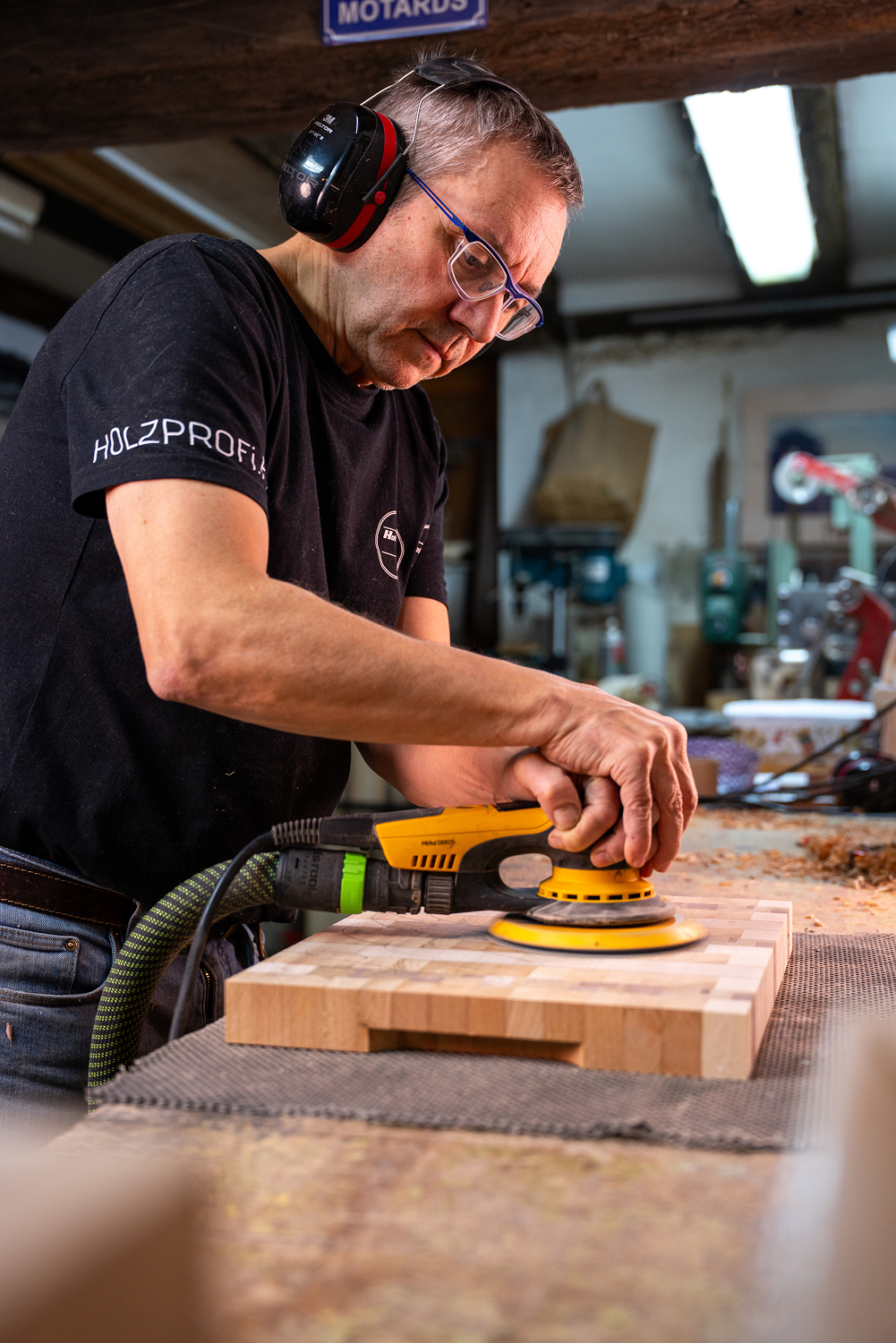 Homme avec lunettes et casque antibruit utilisants une ponceuse électrique jaune sur une planche en bois dans un atelier de menuiserie.
