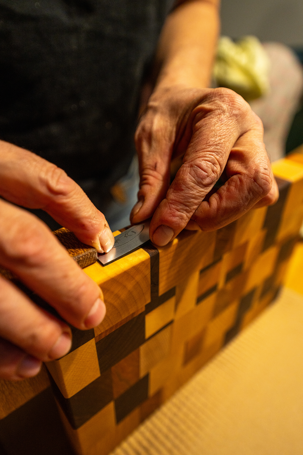Mains d'une personne travaillant sur une planche à découper en bois à motifs carrés avec un petit outil métallique.