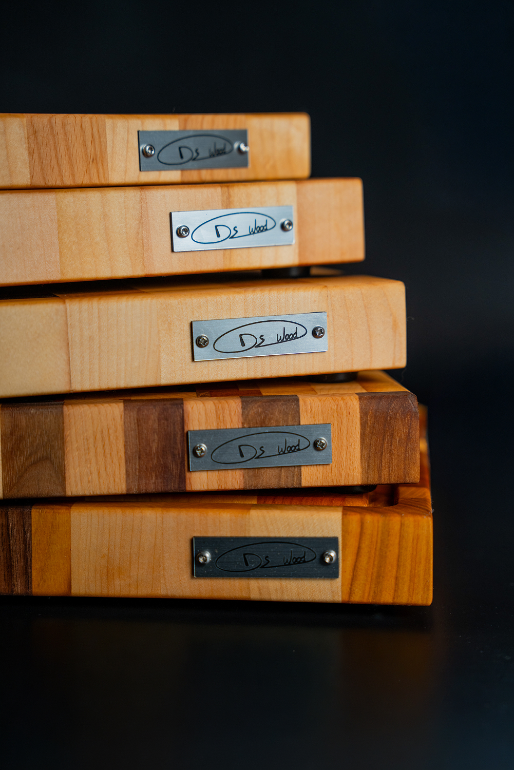 Pile de planches à découper en bois naturel avec plaques métalliques gravées "Ds Wood" sur fond noir.