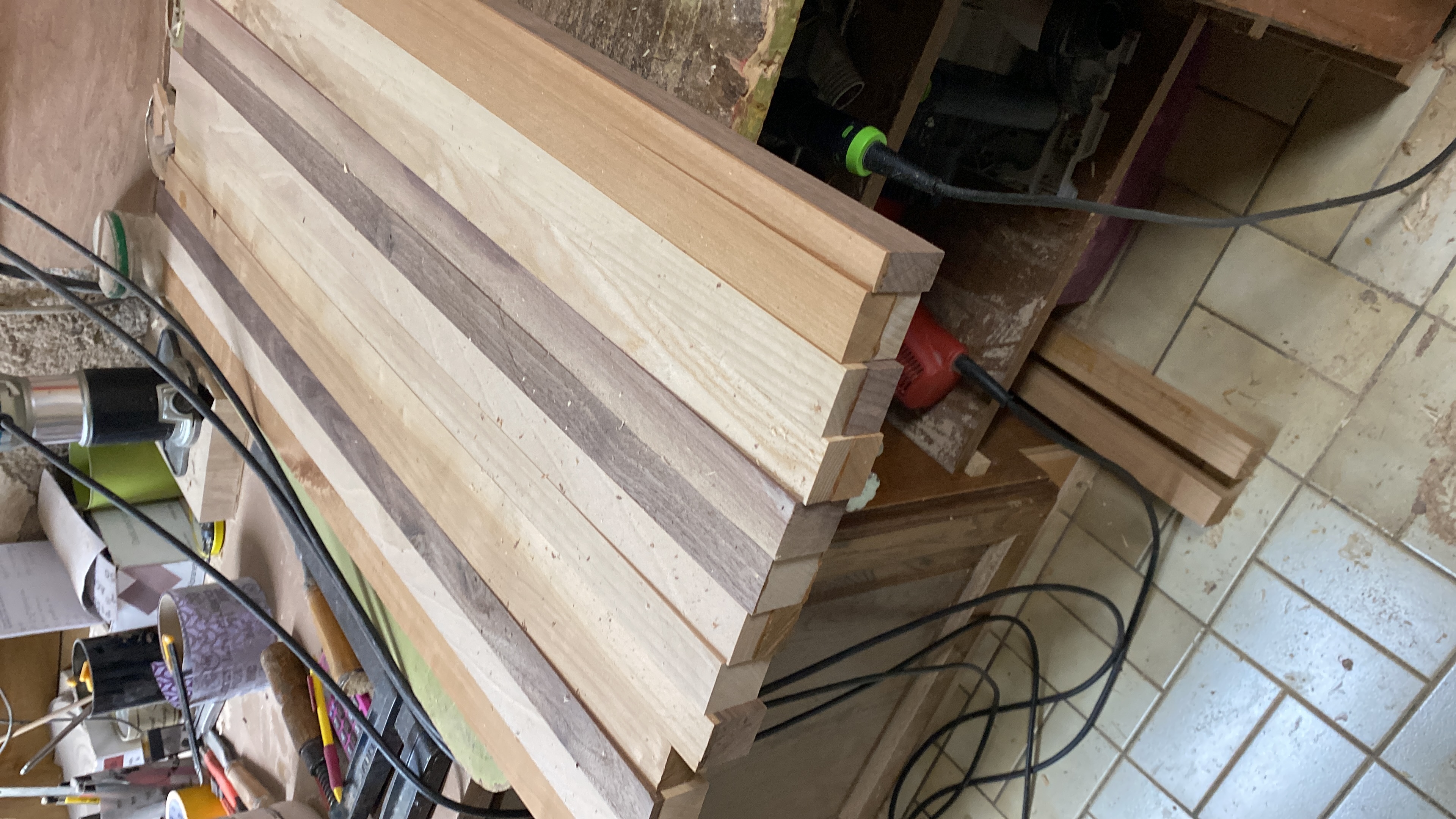 Planches de bois disposées sur un établi dans un atelier de menuiserie avec des outils et câbles électriques visibles.