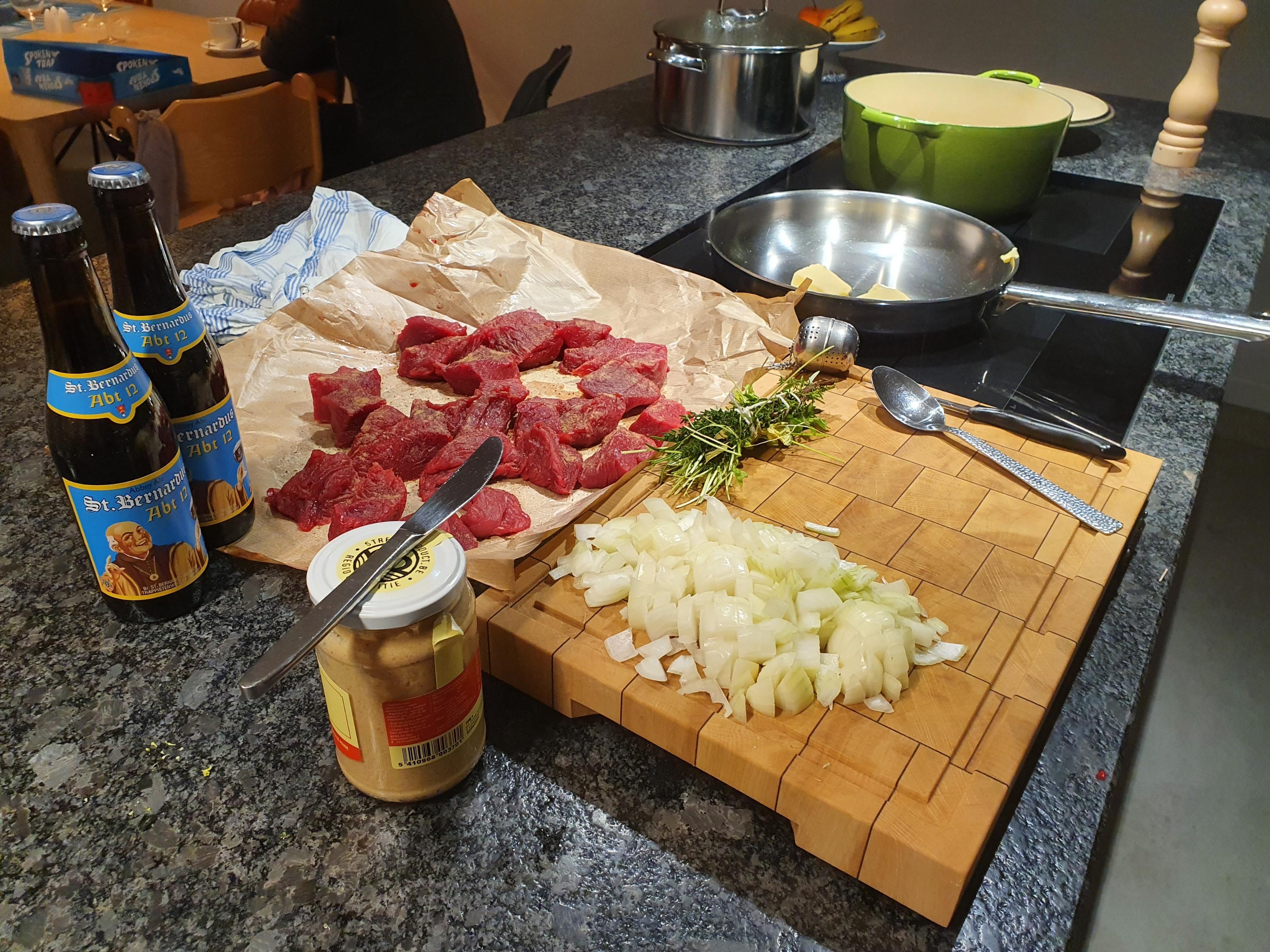 Planche en bois avec viande de bœuf crue, oignons hachés, herbes, moutarde et deux bouteilles de bière, posée sur un plan de travail de cuisine avec une poêle et une cocotte en arrière-plan.