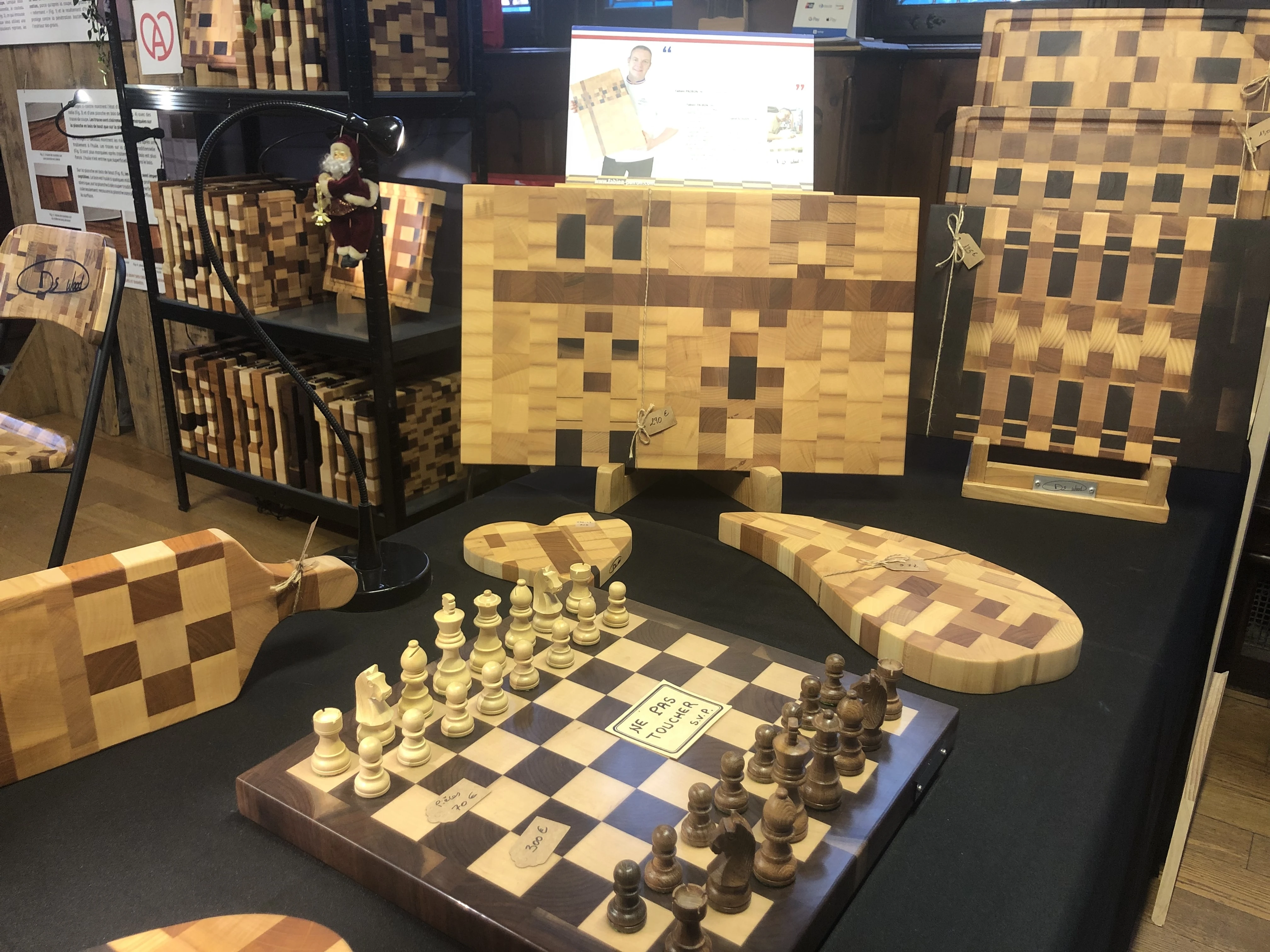 Échiquier en bois avec pièces en place, entouré de planches à découper en bois de différents motifs et tailles exposées sur une table noire.