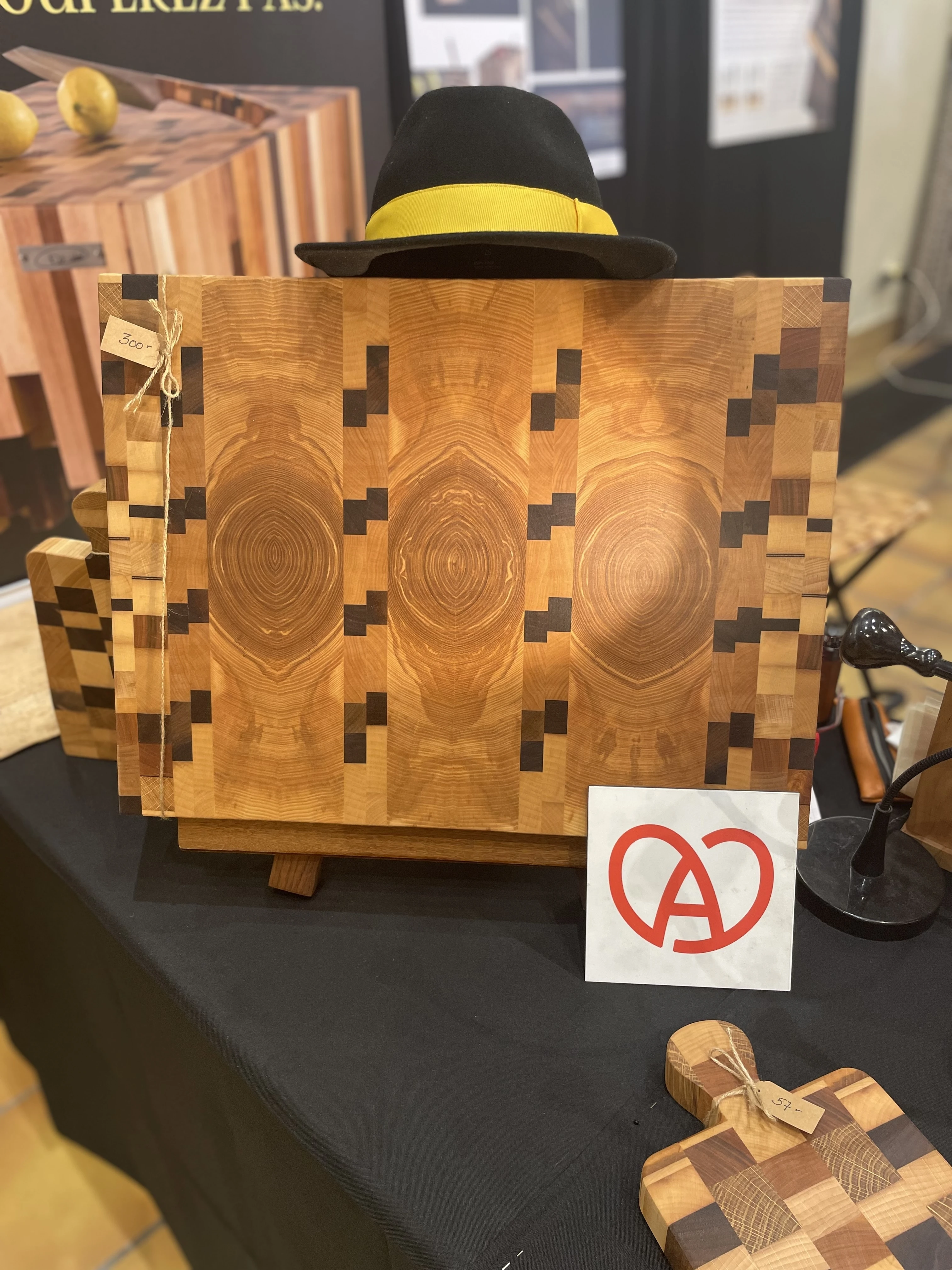 Planche à découper en bois avec motif géométrique, chapeau noir avec bande jaune posé dessus, étiquette de prix et logo rouge sur fond blanc posés à côté.