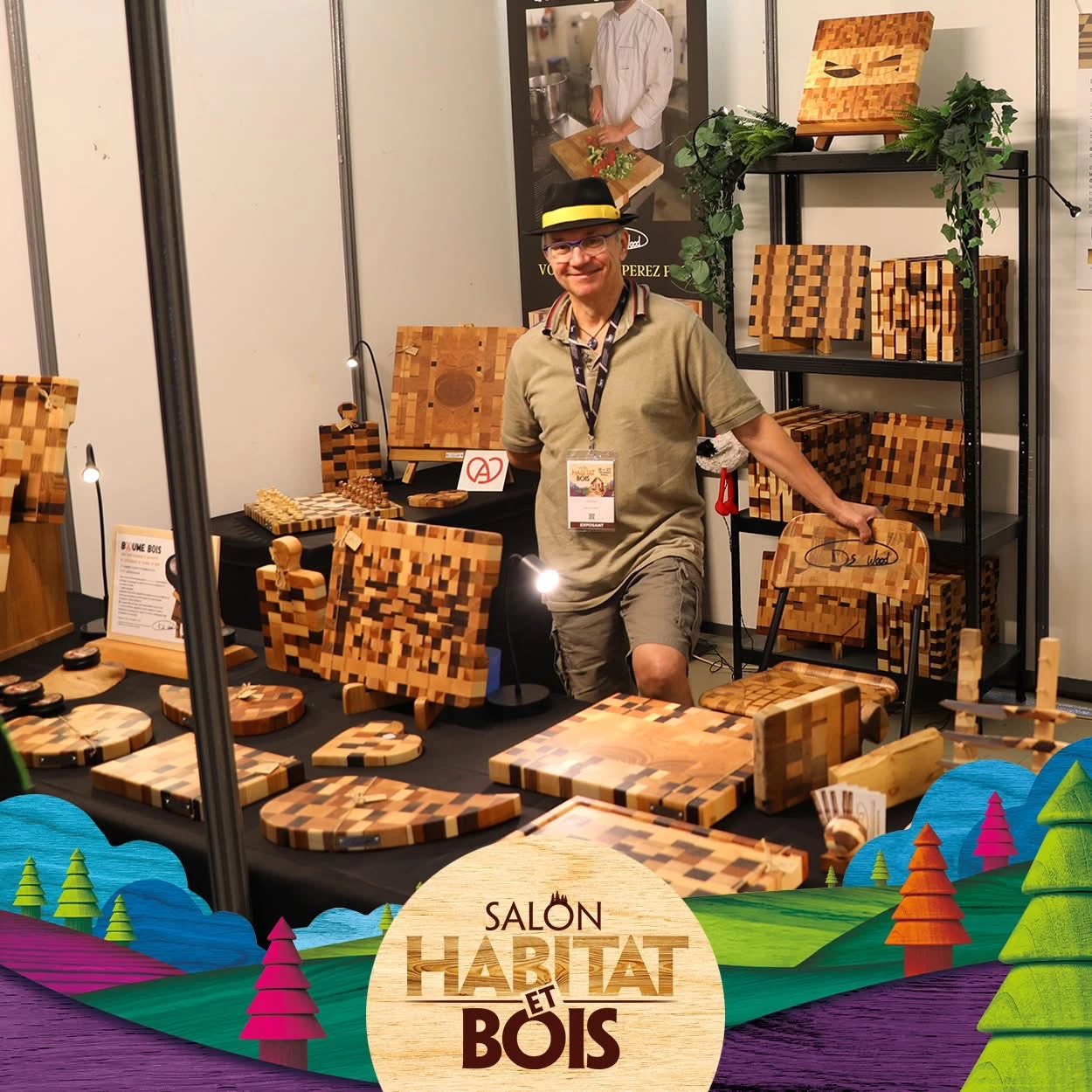 Homme souriant avec un chapeau noir à bande jaune debout à un stand d'exposition présentant des objets en bois aux motifs carrés, dont des planches, un jeu d'échecs et des décorations, au Salon Habitat et Bois.