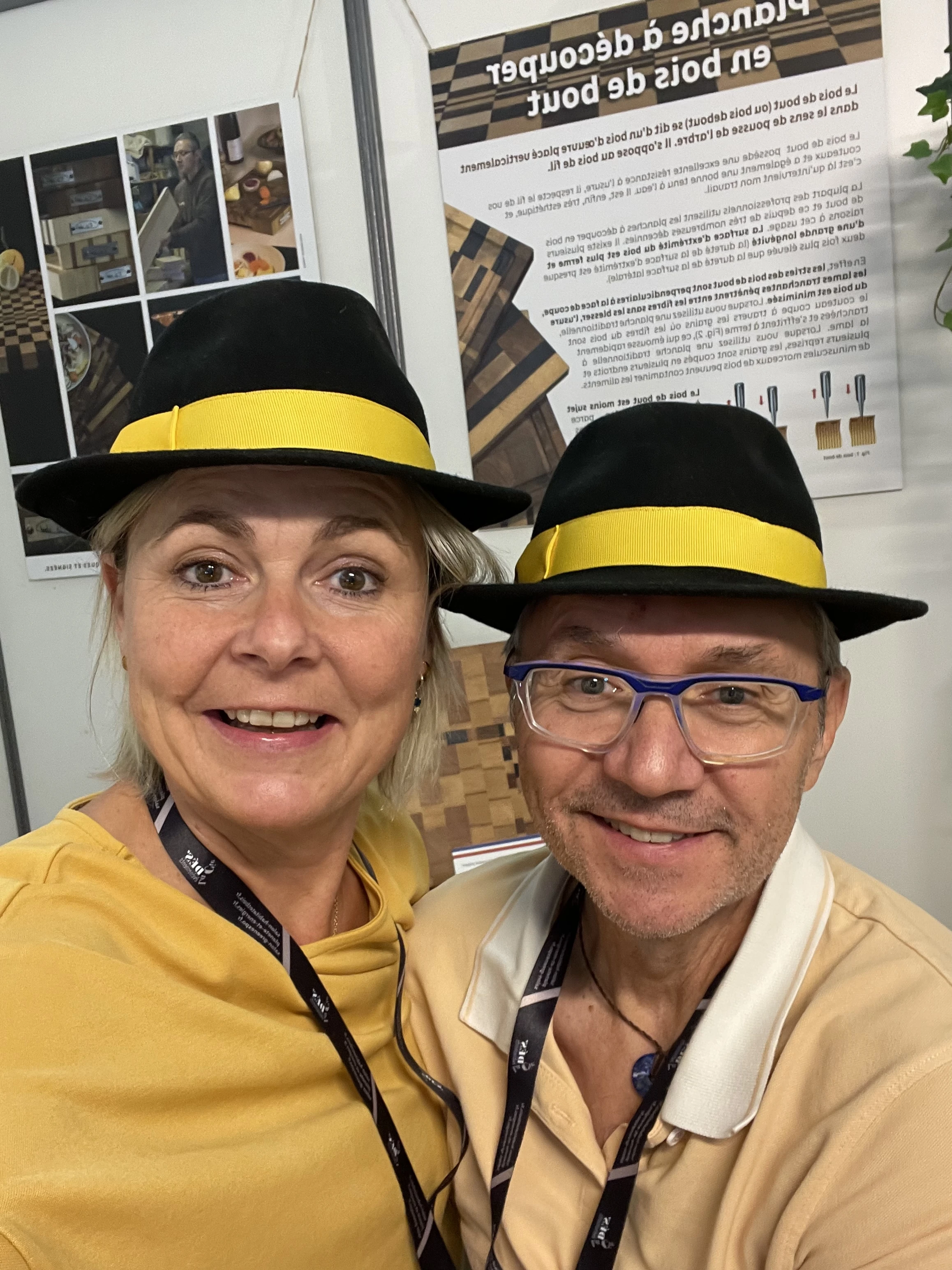 Portrait de deux personnes souriantes portant des chapeaux noirs à bande jaune, avec des panneaux d'exposition en arrière-plan.