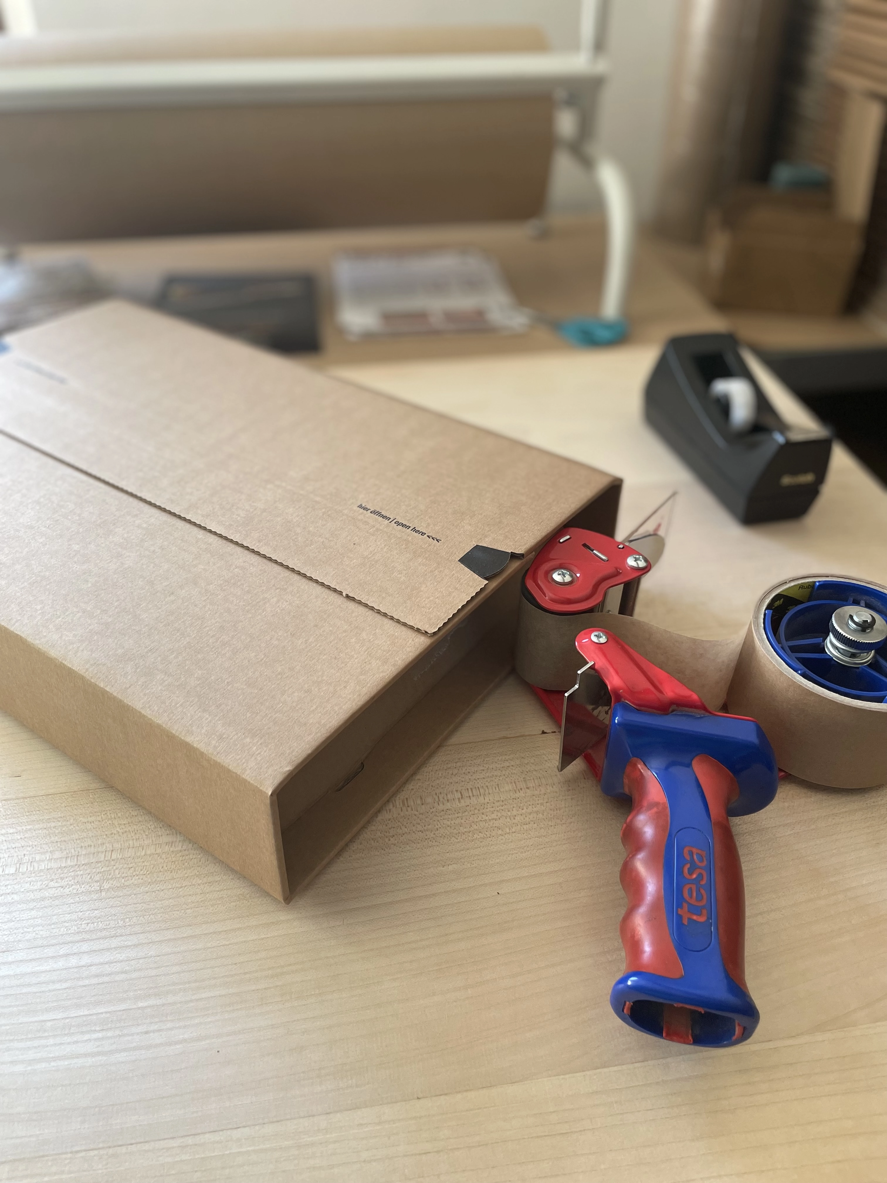Un ruban adhésif marron monté sur un dévidoir bleu et rouge marqué 'tesa', à côté d'une grande boîte en carton sur une table en bois.