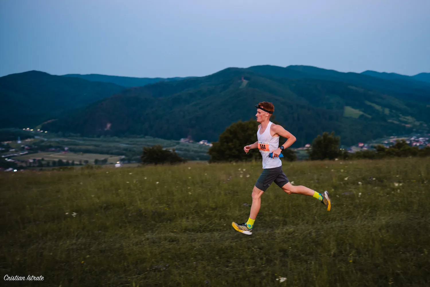 Young male runner wearing a white tank top, black shorts, and yellow socks running on a grassy field with mountains in the background.