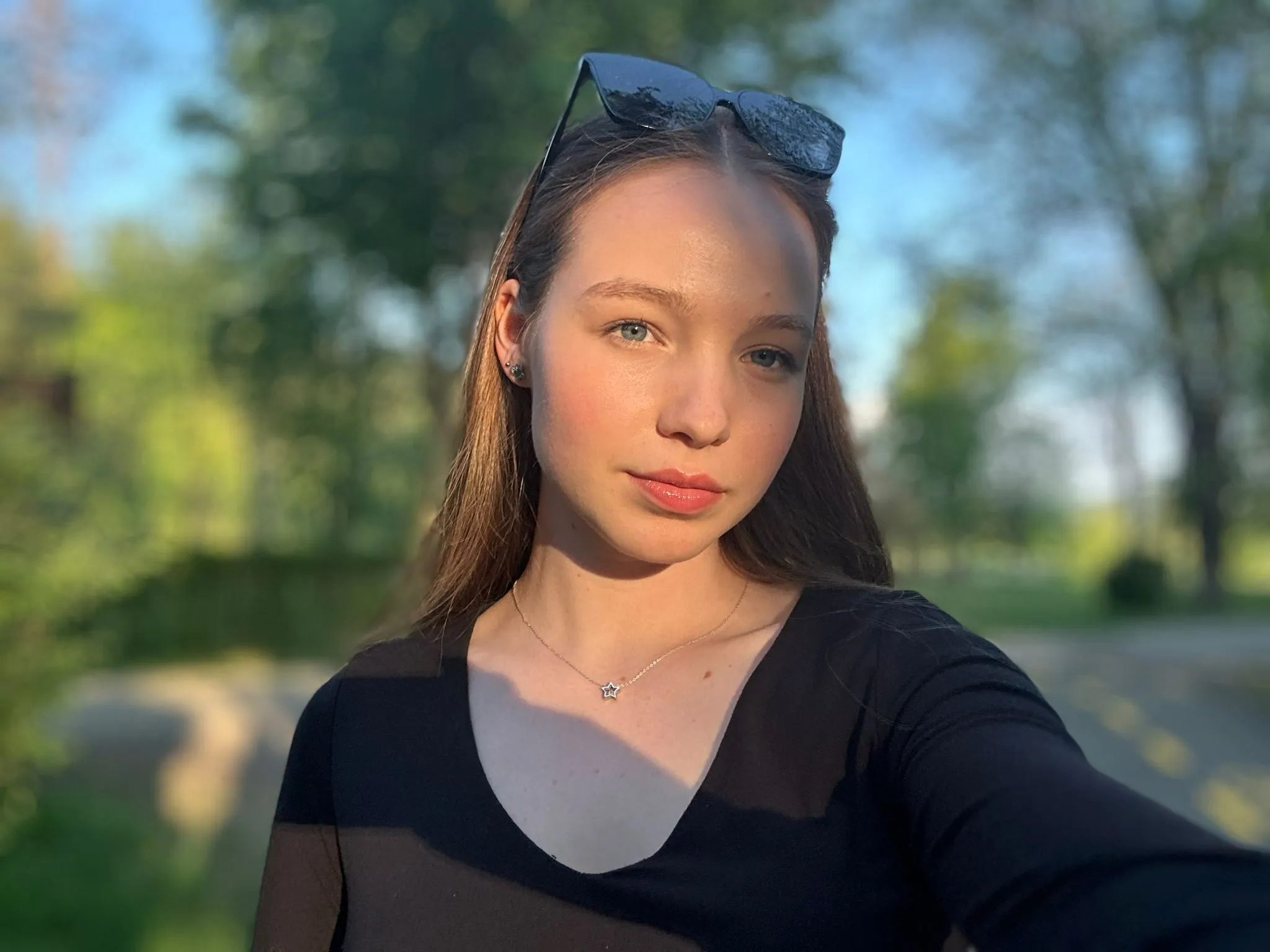 Young woman with long hair wearing black top and star necklace, sunglasses on head, outdoors with blurred green trees background.