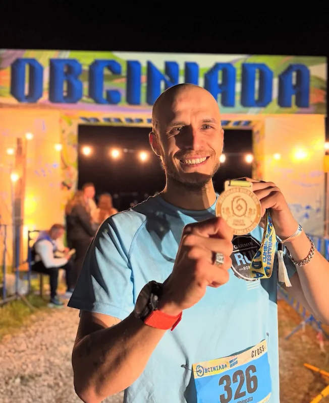 Smiling bald man in a light blue running shirt holding a finisher medal in front of a brightly lit race finish banner reading 'OBCINIADA'.