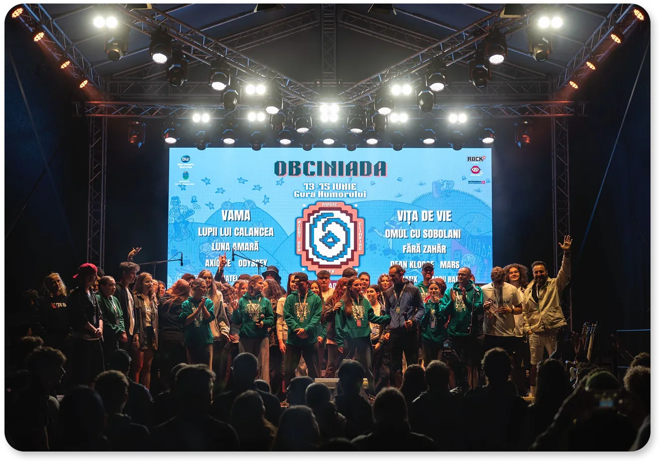 Group of people standing on a brightly lit stage in front of a large screen displaying 'Obciniada' festival lineup and dates.