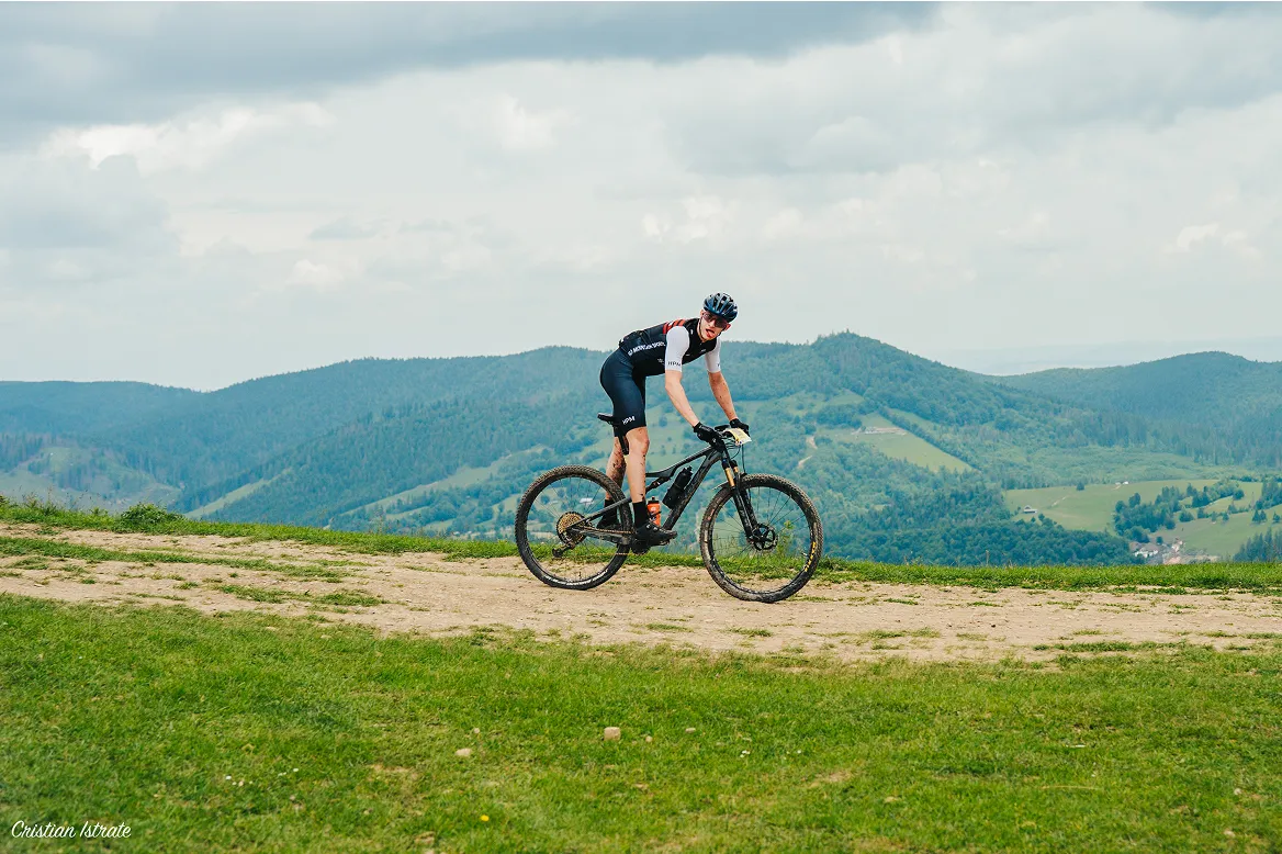 Cyclist in black gear riding a mountain bike on a dirt path with green hills and cloudy sky in the background.