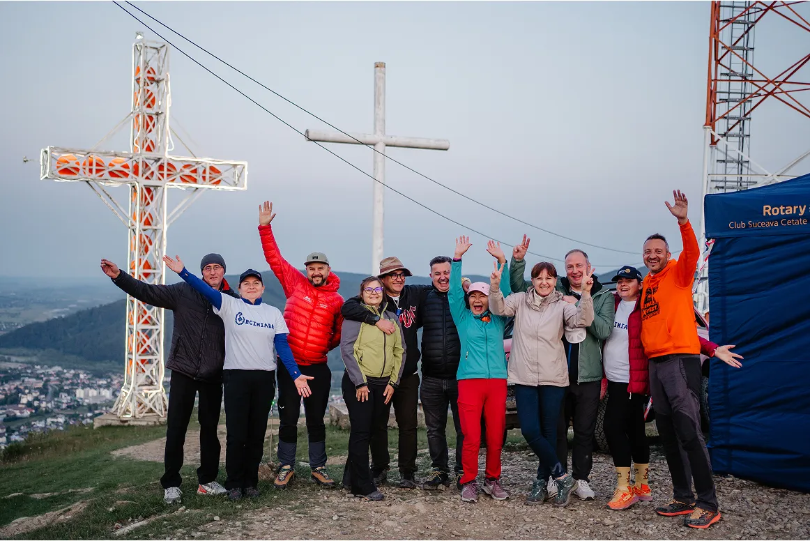 Group of eleven people standing on a hilltop with arms raised, smiling in front of two large crosses and a Rotary Club tent at dusk.