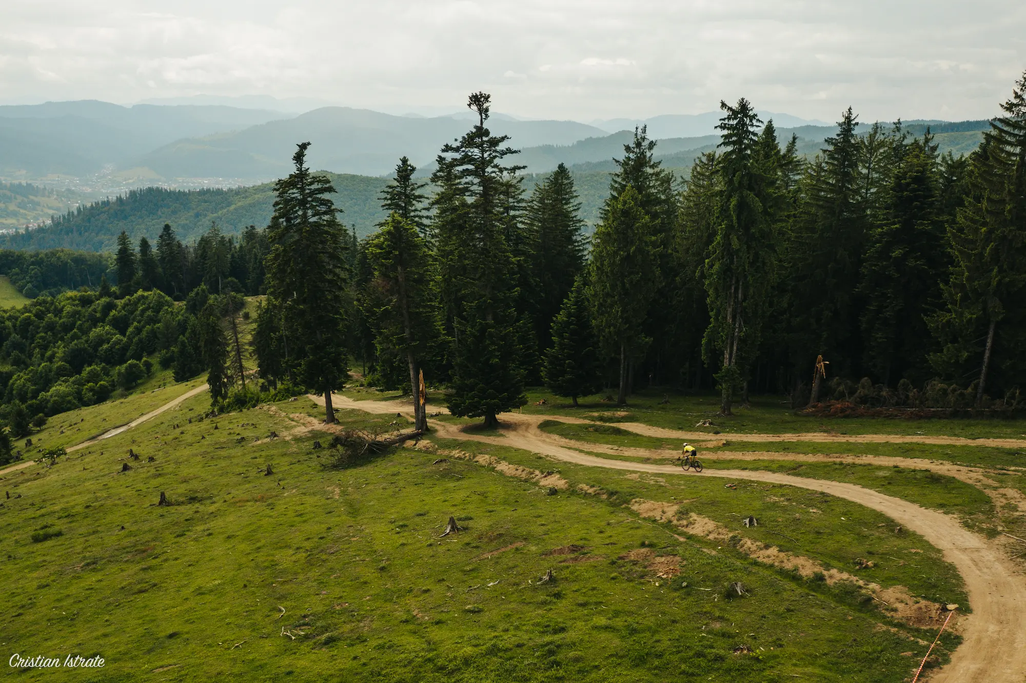 Cyclist riding on a winding dirt path through a green forested hillside with mountains in the distance under a cloudy sky.