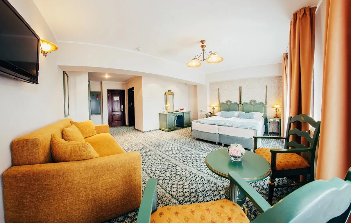 Bright hotel room with a mustard yellow sofa, green wooden furniture, twin beds, and orange curtains.