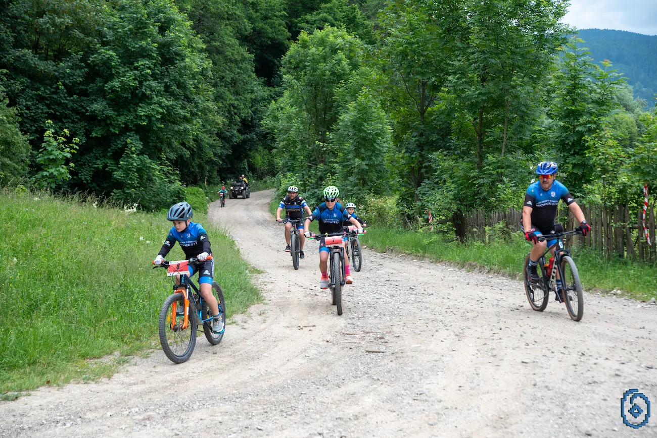 Group of cyclists, including children and an adult, riding on a gravel path through a green, wooded area.