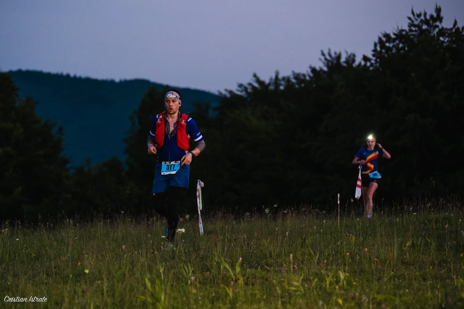 Two runners participating in a trail race at dusk, one wearing a headlamp and the other in a red vest, running through a grassy field with forest and hills in the background.