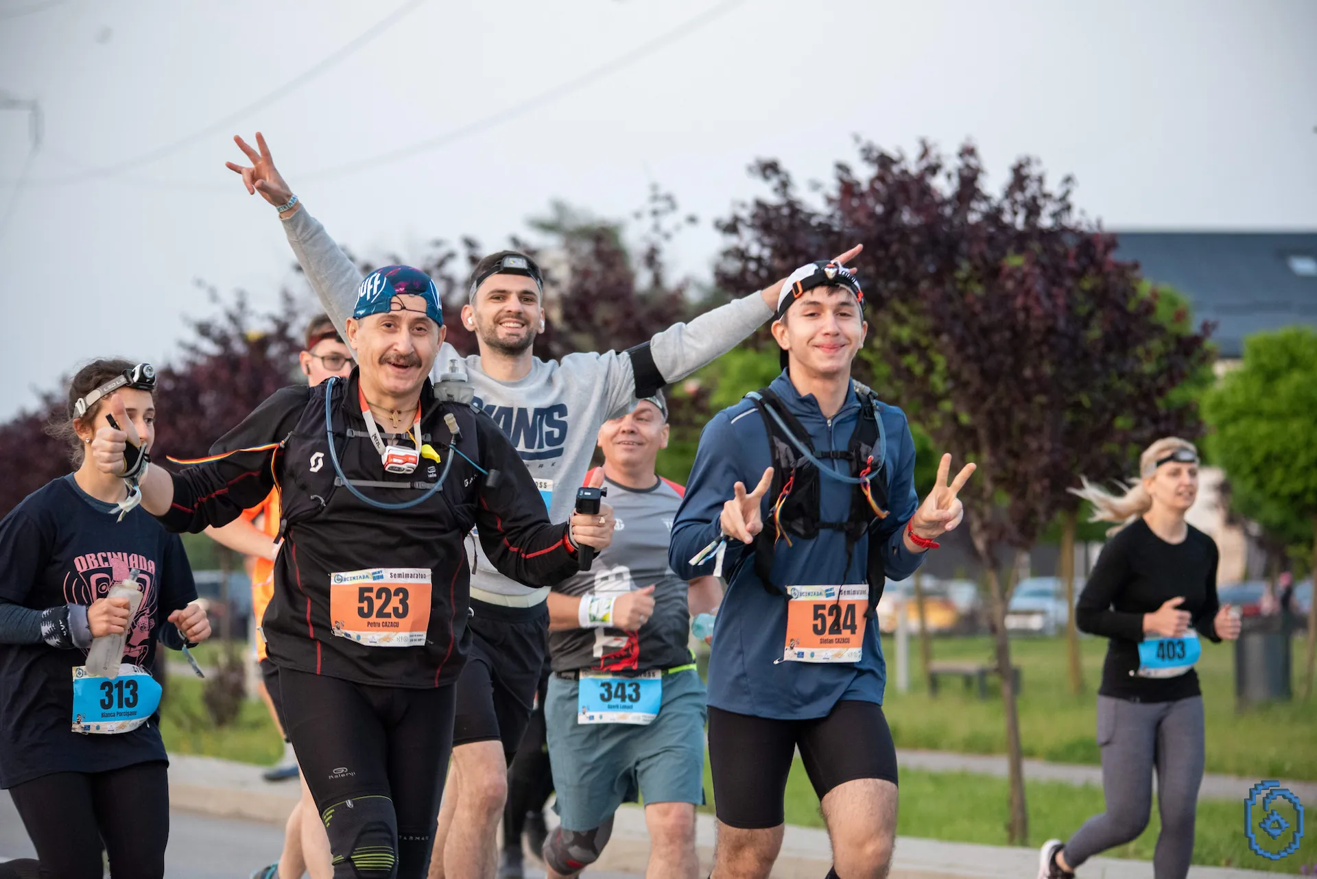 Group of runners smiling and making peace signs during an outdoor race on a paved path with trees in the background.