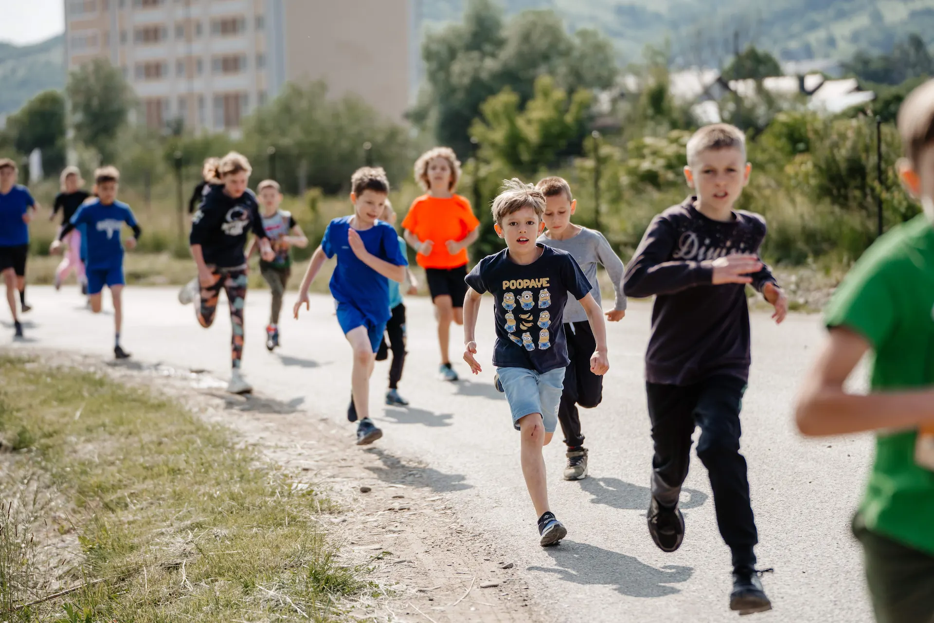 A group of children running outdoors on a paved path with trees and buildings in the background.