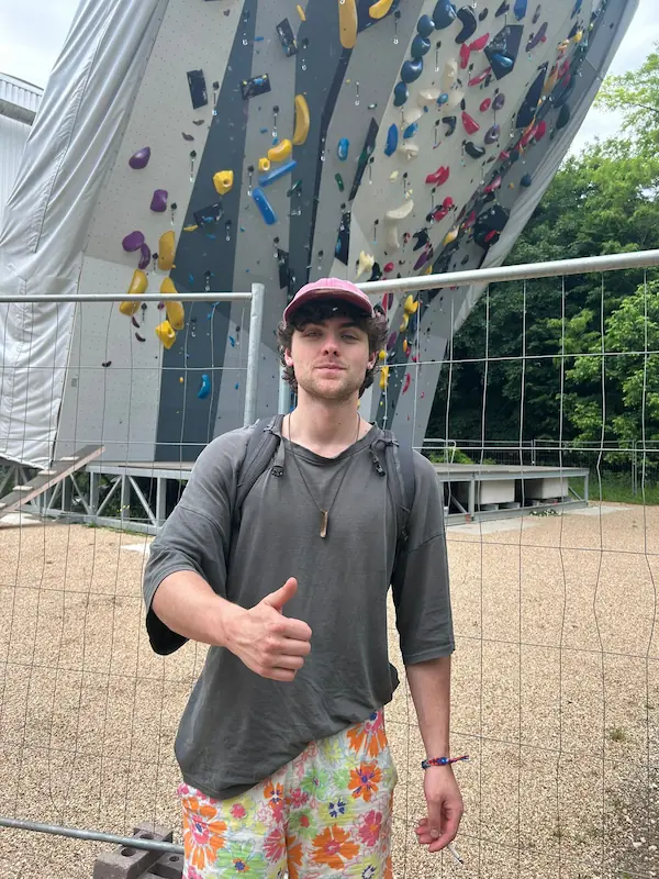 Bărbat tânăr cu șapcă roz, tricou gri și pantaloni scurți cu flori colorate făcând semn de bine în fața unui perete de escaladă cu prize colorate.