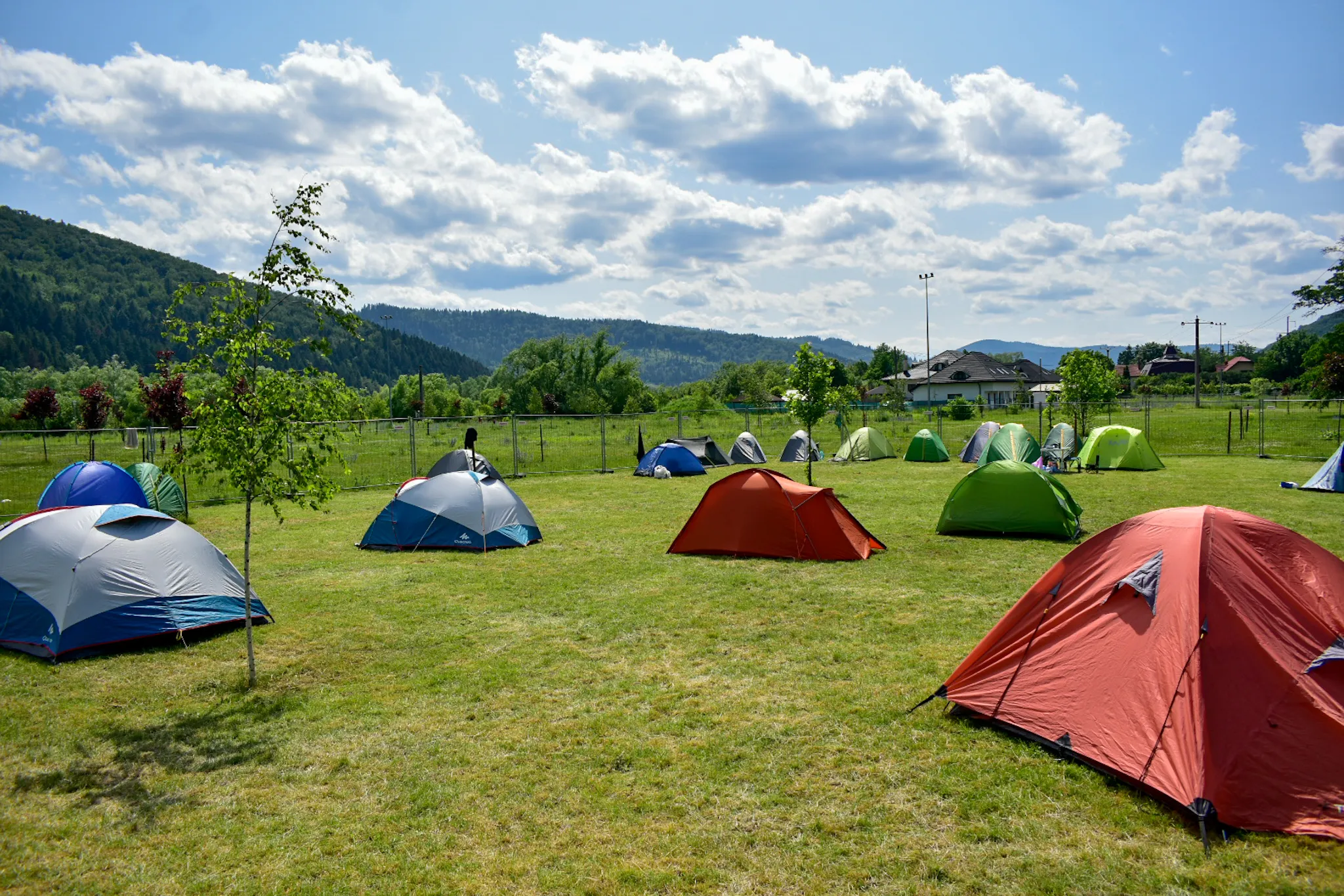 Camping cu corturi viu colorate pe un câmp verde cu dealuri împădurite în fundal sub un cer parțial înnorat.