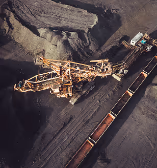 Aerial view of a large industrial excavator loading dark coal or mineral material into open train wagons at a mining site, surrounded by piles of raw material and dusty ground in low sunlight.