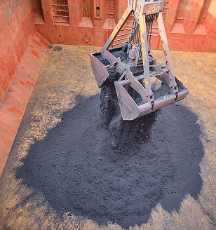 Close-up of a metal grab crane unloading dark coal or mineral cargo into the hold of a large ship, forming a pile on the rusty floor of the vessel’s interior storage compartment.
