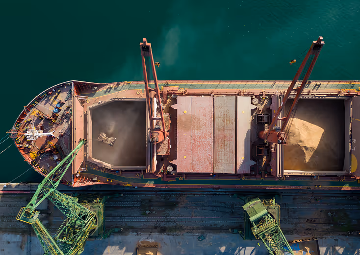 Aerial view of a bulk cargo ship docked at a port being loaded with grain or sand using large green cranes, with cargo holds open and dust rising as material is poured into the compartments over calm teal water.