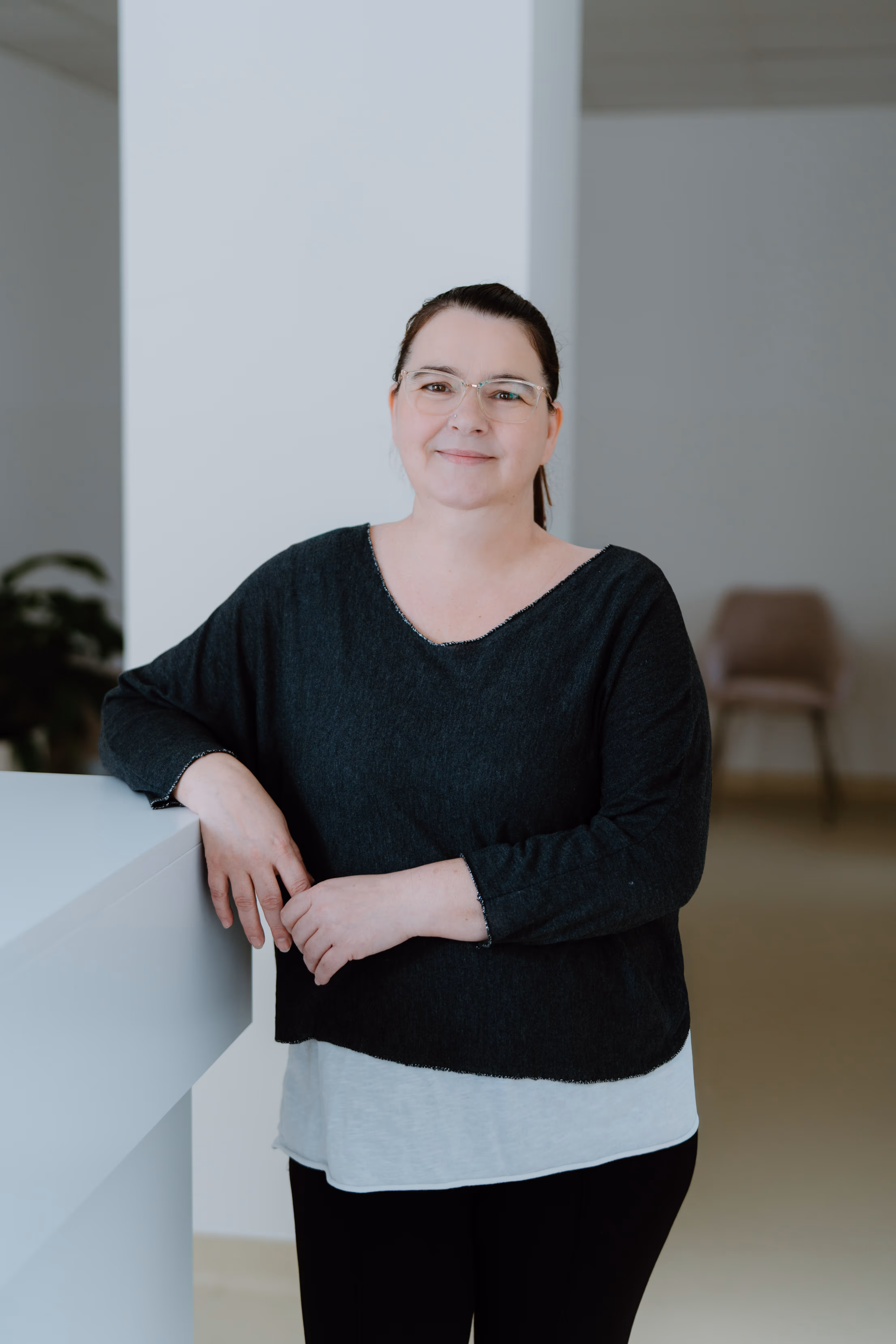Frau mit Brille und schwarzem Pullover lehnt lächelnd an einer weißen Theke in einem modernen Innenraum.