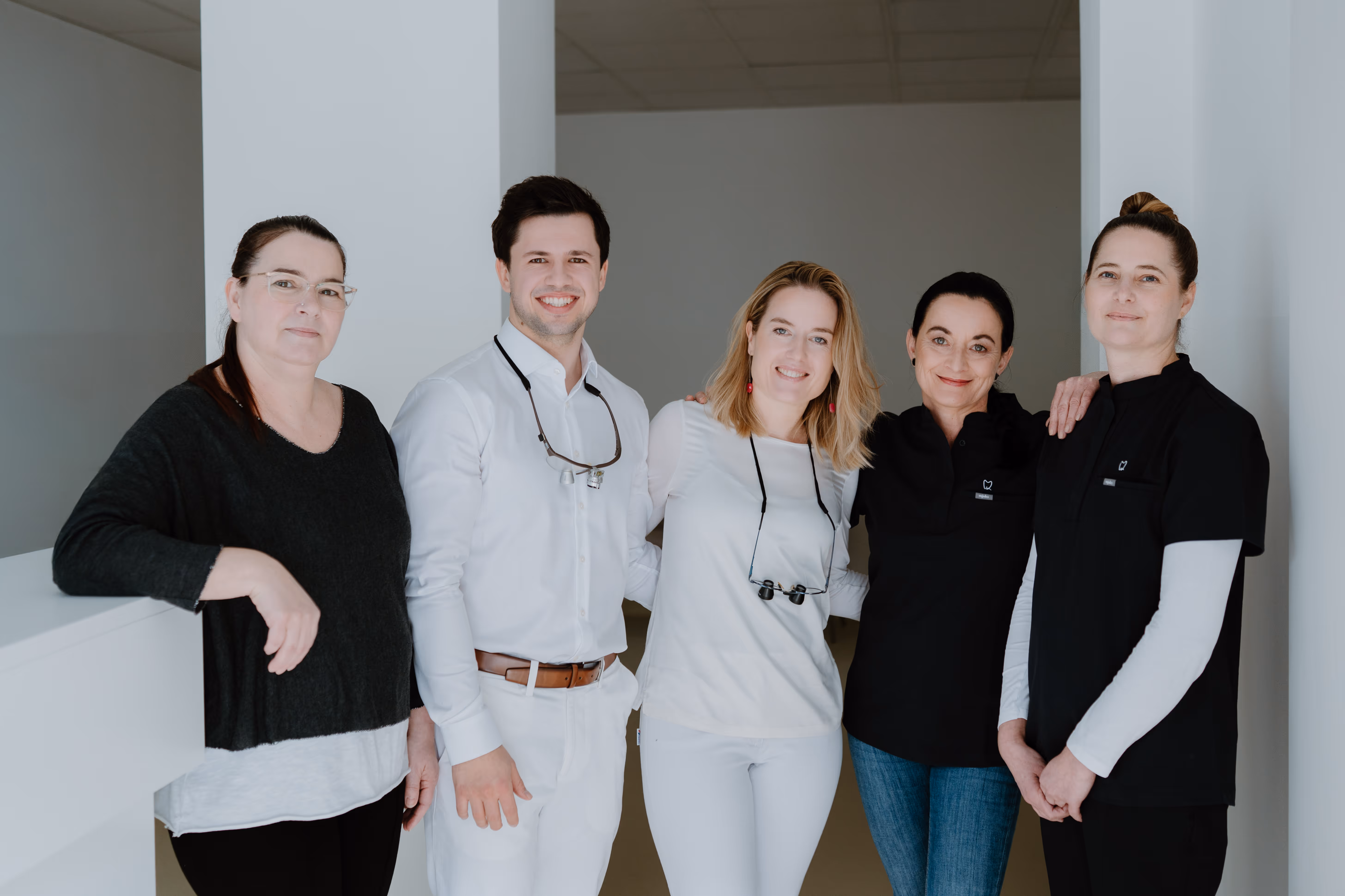 Fünf Mitarbeiter eines Zahnarztteams stehen nebeneinander in einer Praxis und lächeln in die Kamera.