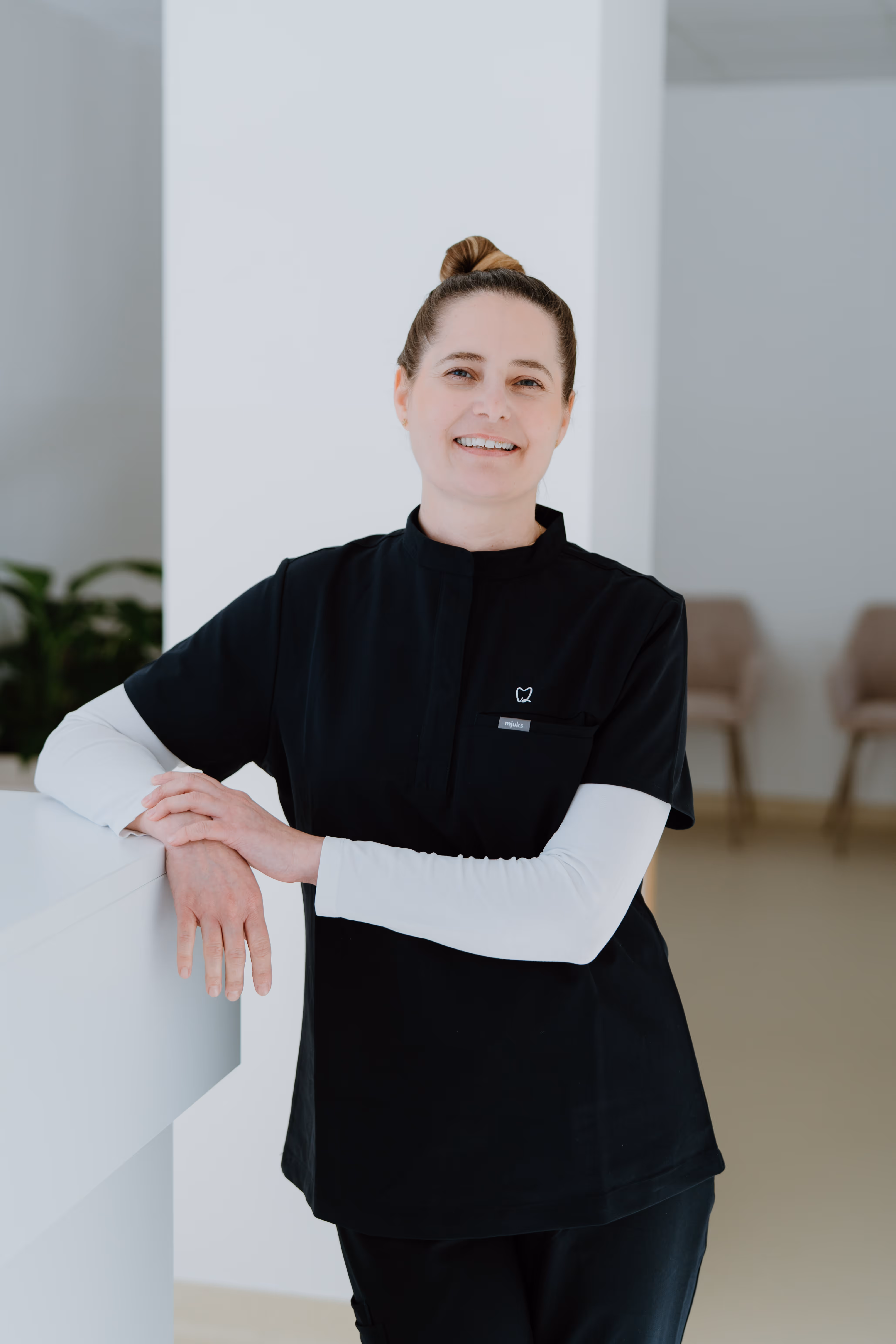 Lächelnde Frau in schwarzer Zahnarzt- oder medizinischer Arbeitskleidung lehnt an einem weißen Empfangstresen.