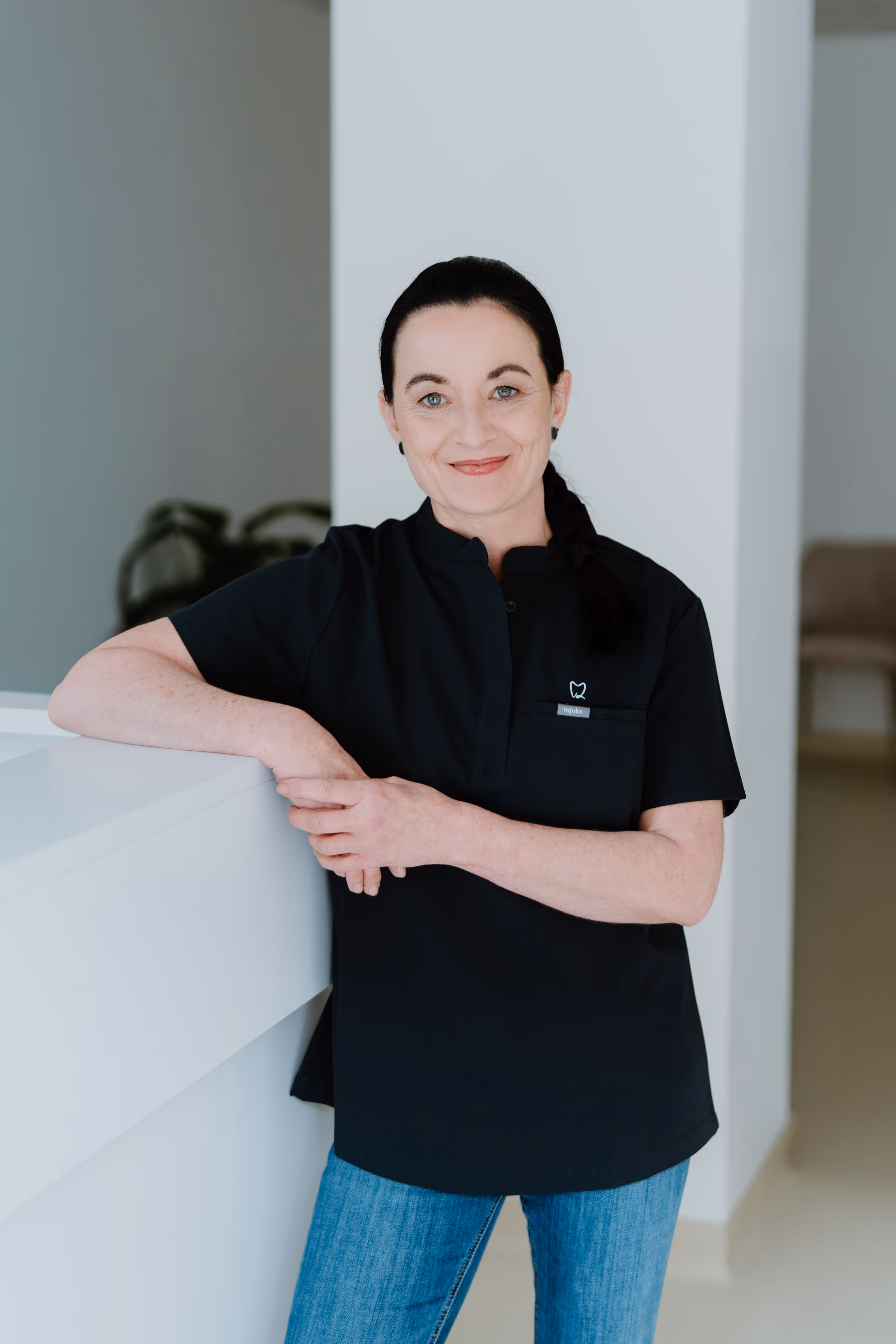 Eine lächelnde Frau mit dunklen Haaren, schwarzem Oberteil mit Zahn-Logo und blauen Jeans lehnt an einem weißen Tresen.