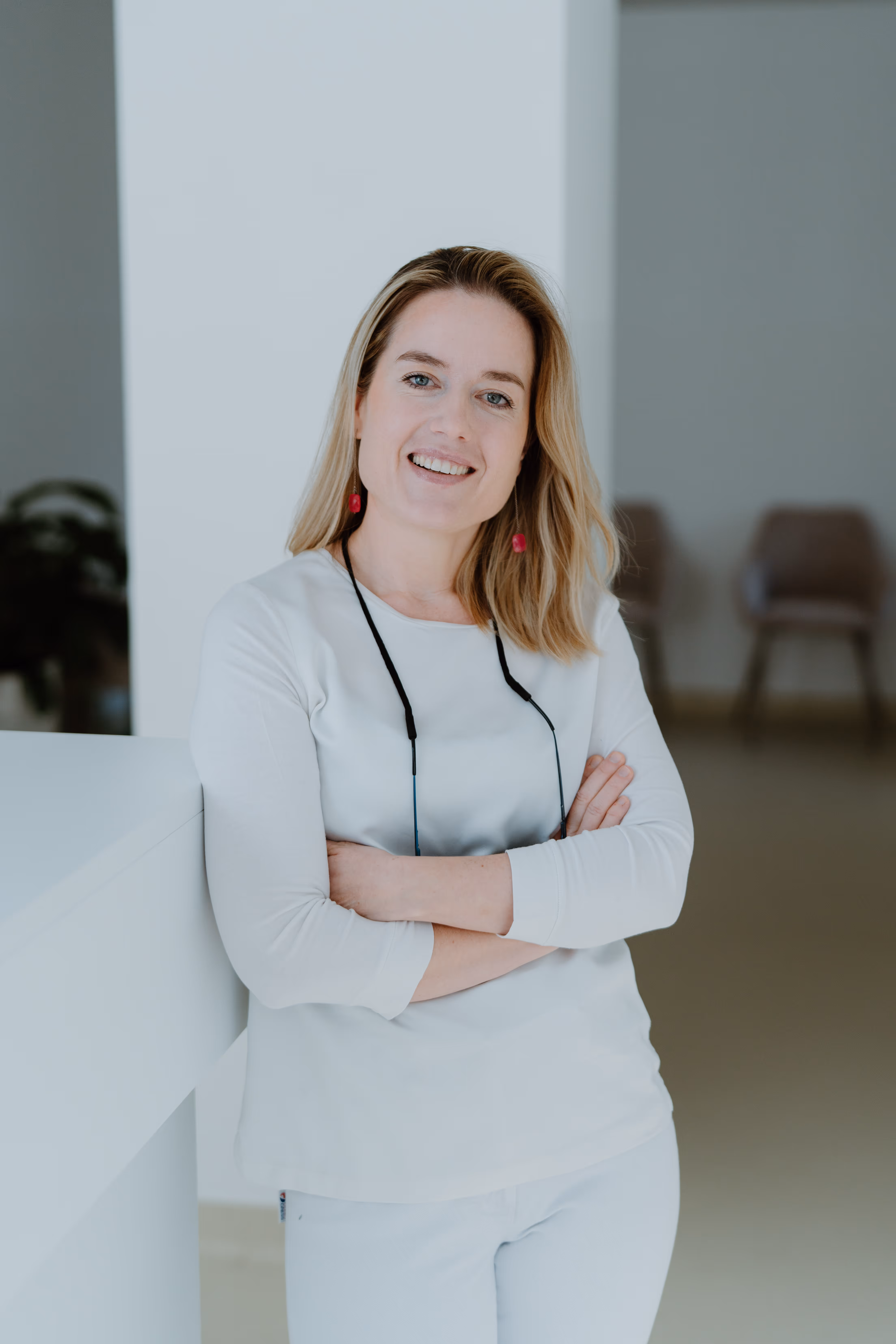 Lächelnde Frau mit verschränkten Armen lehnt an einem weißen Tisch in einem hellen Raum.