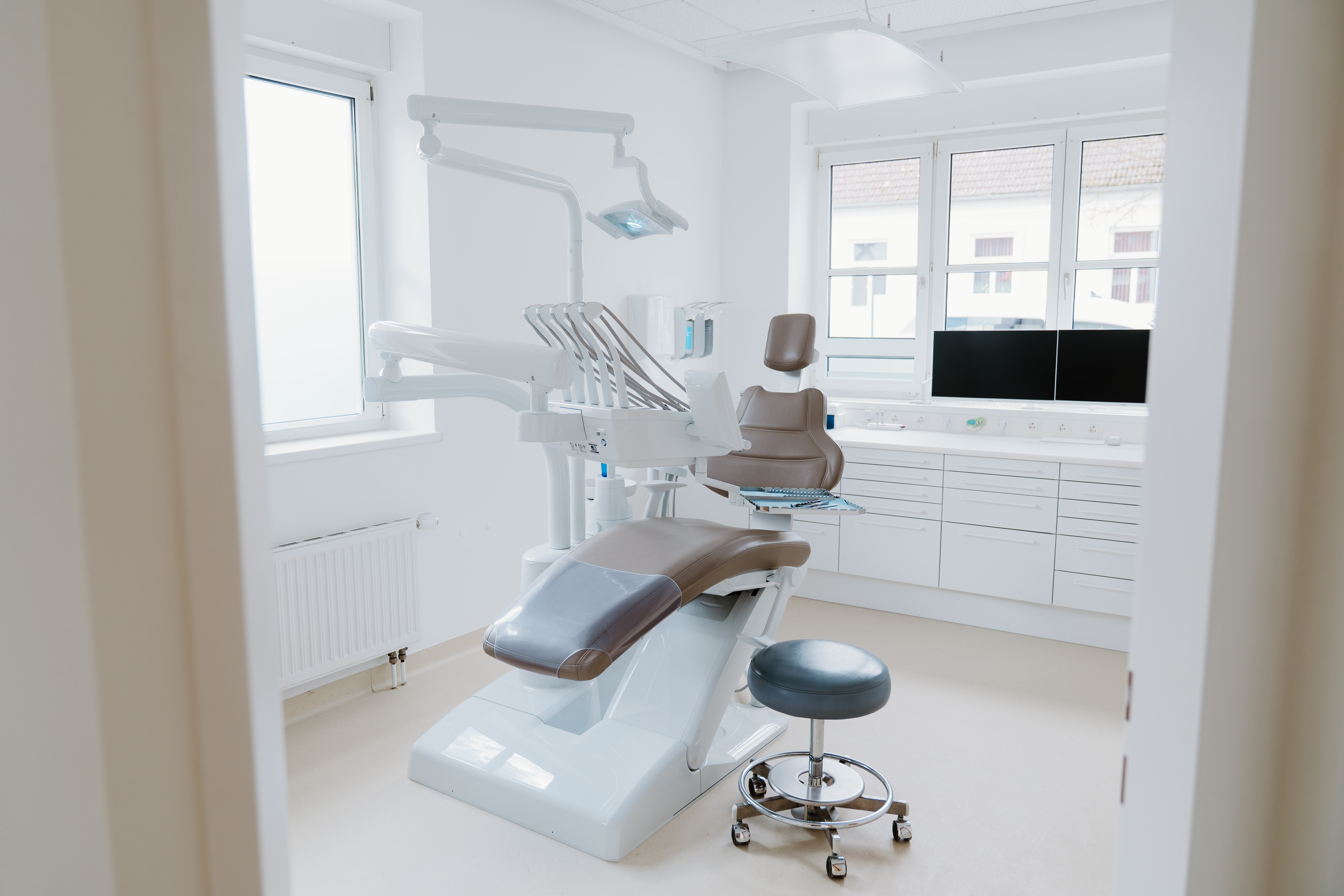 Moderne Zahnarztpraxis mit einem braunen Behandlungsstuhl, Instrumentenarm und Hocker vor Fenstern mit Tageslicht.