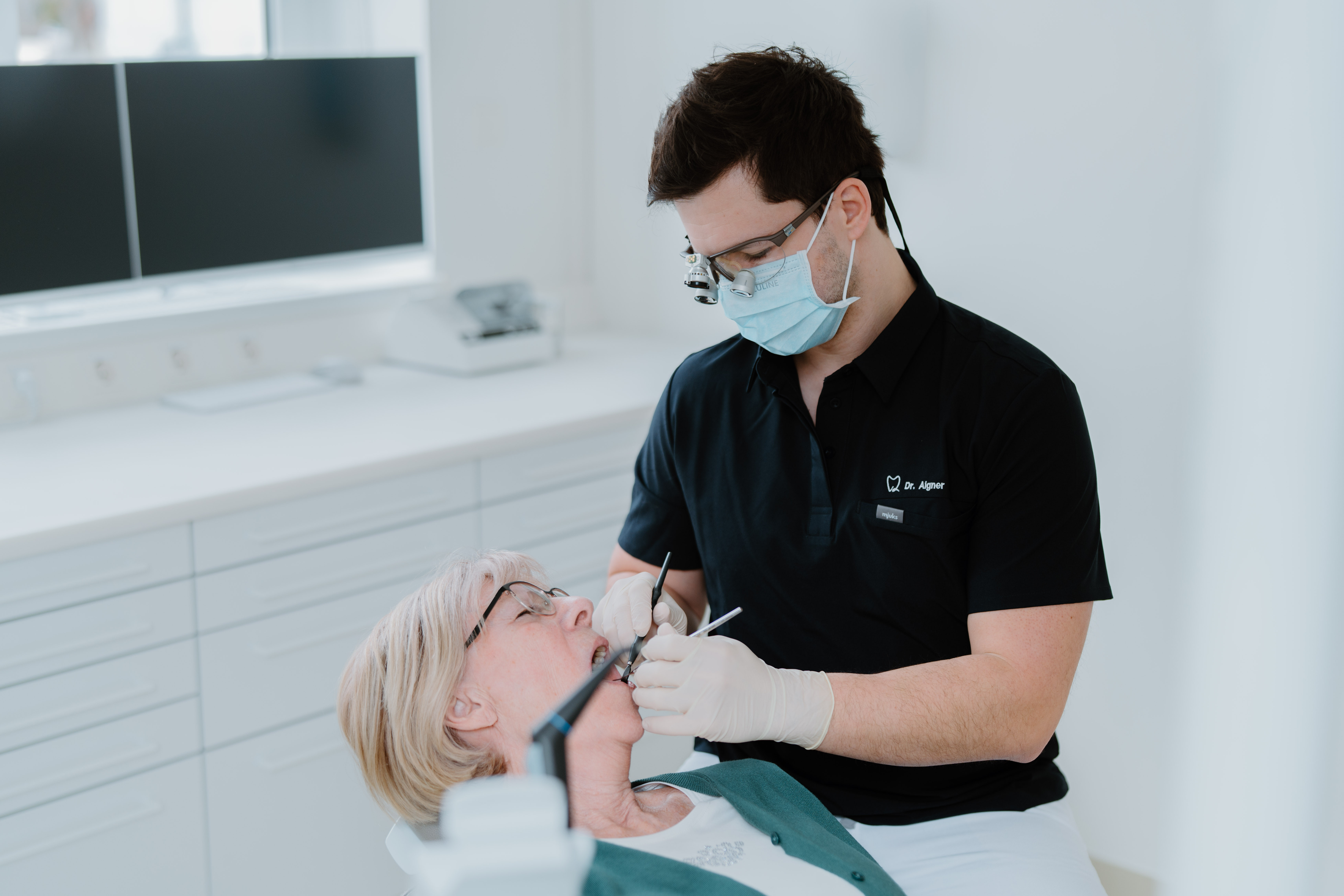 Zahnarzt mit Mundschutz und Lupenbrille behandelt ältere Patientin in heller Zahnarztpraxis.
