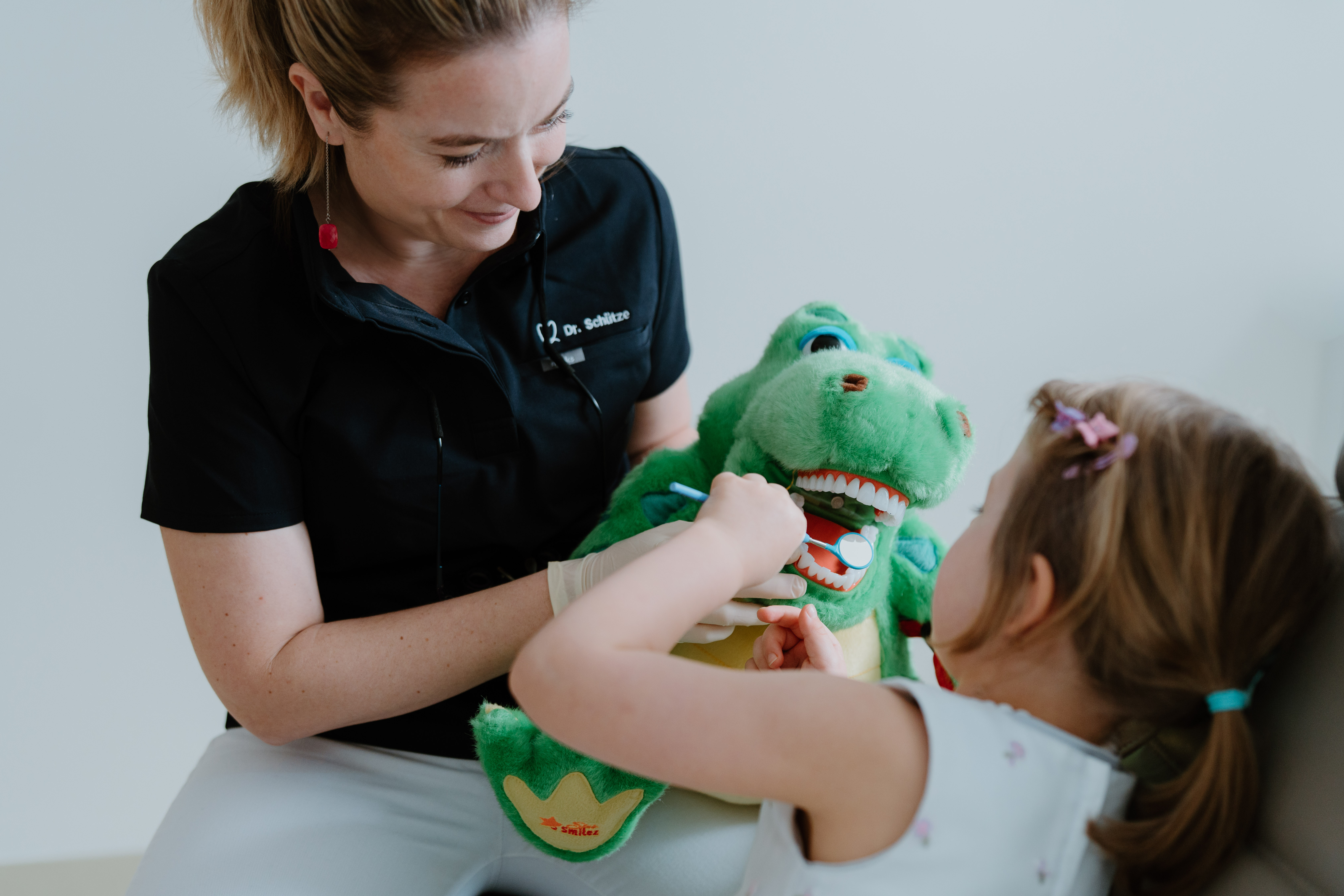 Zahnärztin erklärt einem kleinen Mädchen an einem grünen Krokodil-Plüschtier die Zahnpflege.