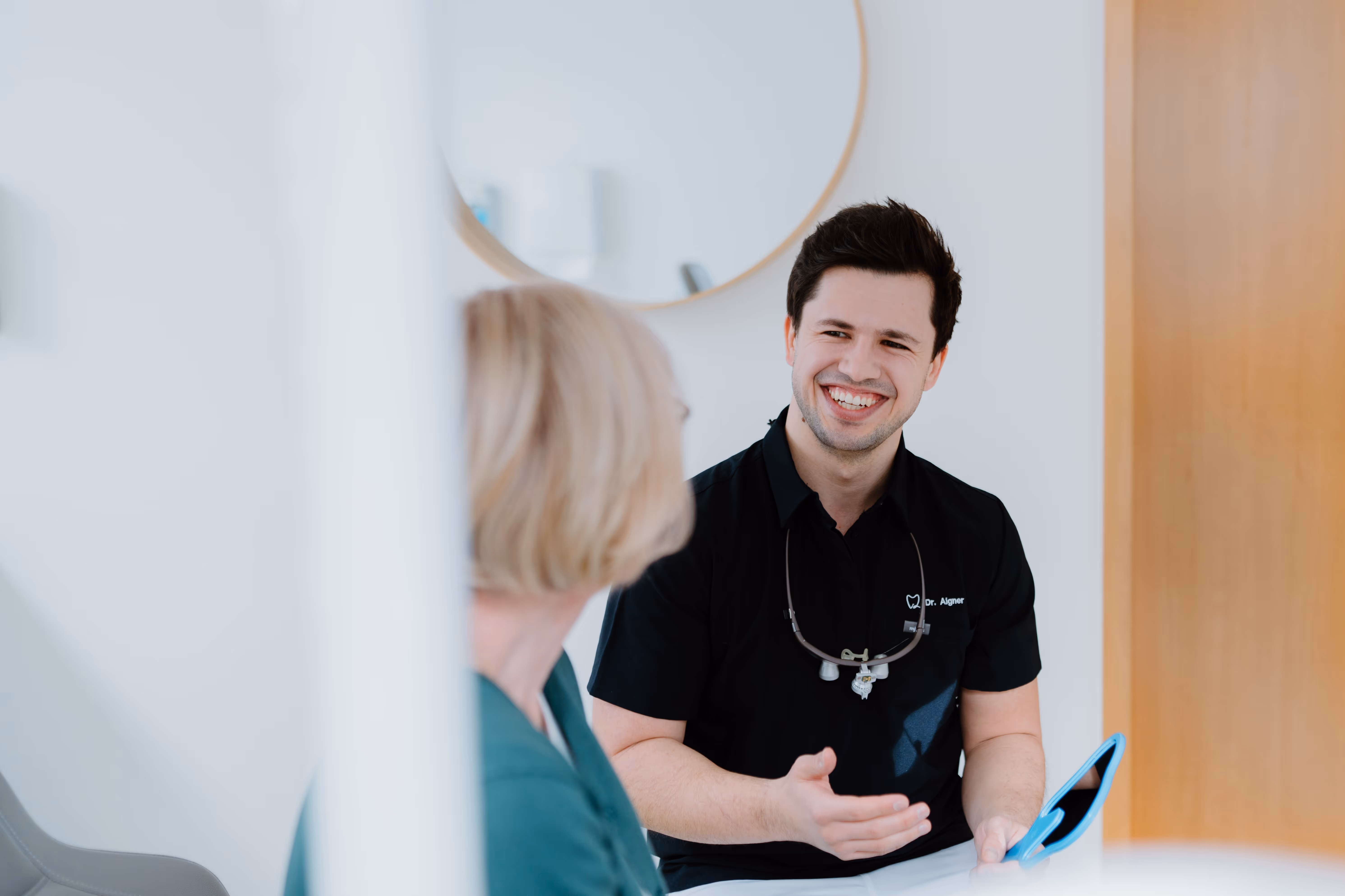 Zahnarzt im Gespräch mit Patientin in heller Praxis, er hält einen Zahnspiegel.