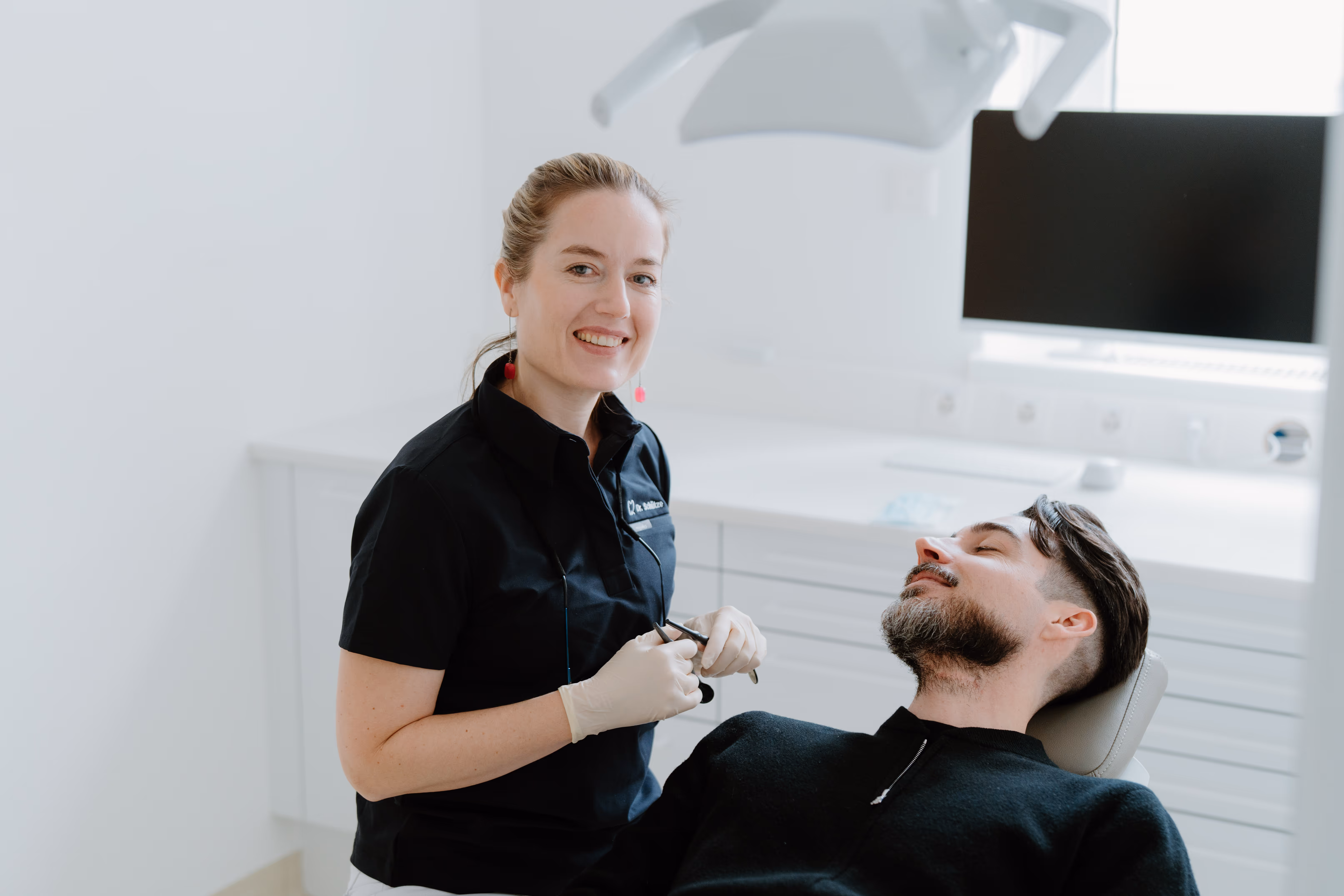 Zahnärztin lächelt in der Zahnarztpraxis neben einem entspannten Patienten im Behandlungsstuhl.