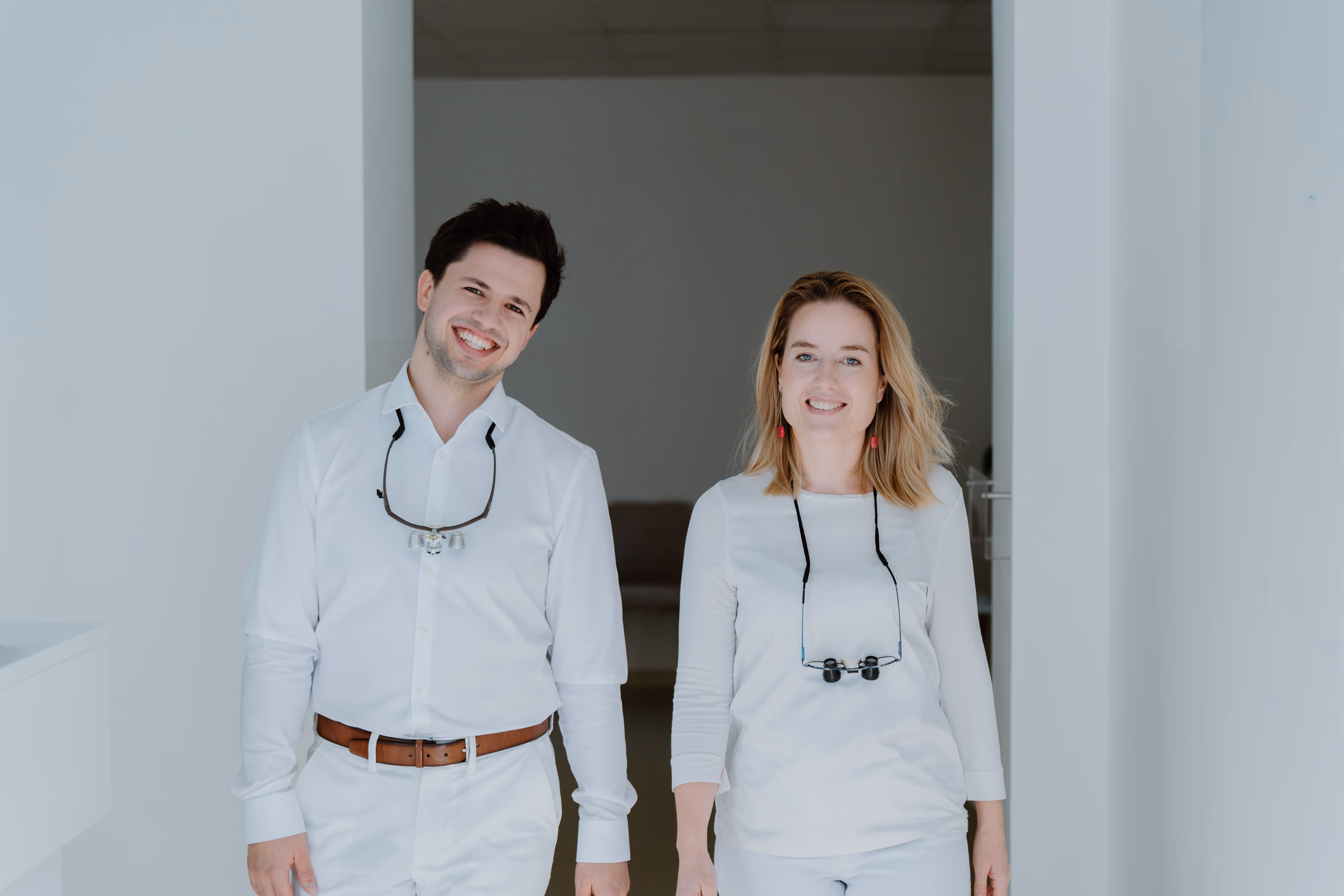 Lächelnder Mann und Frau in weißer Arbeitskleidung mit Lupenbrillen um den Hals in einem hellen Raum.