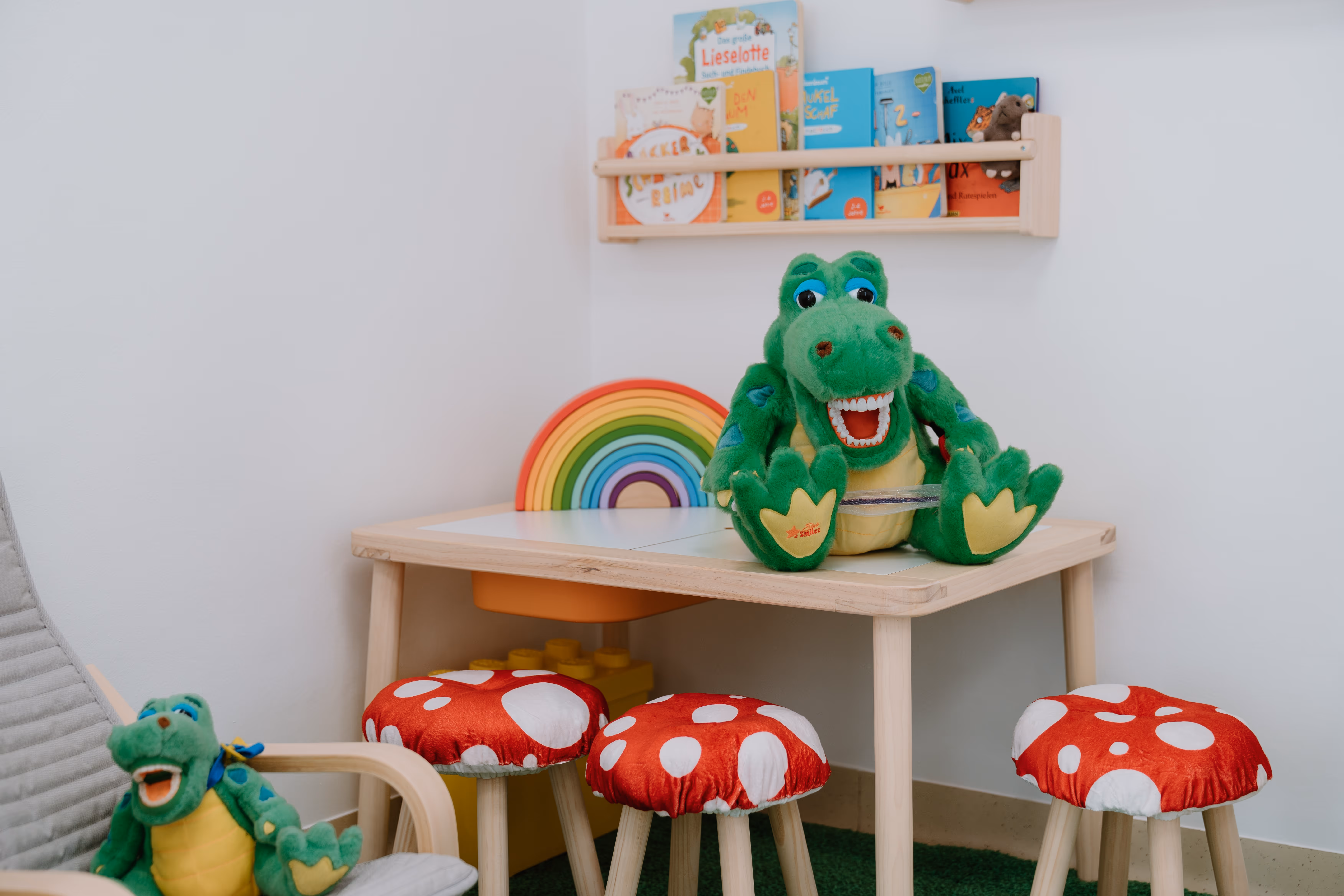 Kinder-Wartebereich mit kleinem Holztisch, buntem Regenbogen-Spielzeug, drei Pilz-Hockern und zwei grünen Krokodil-Stofftieren.
