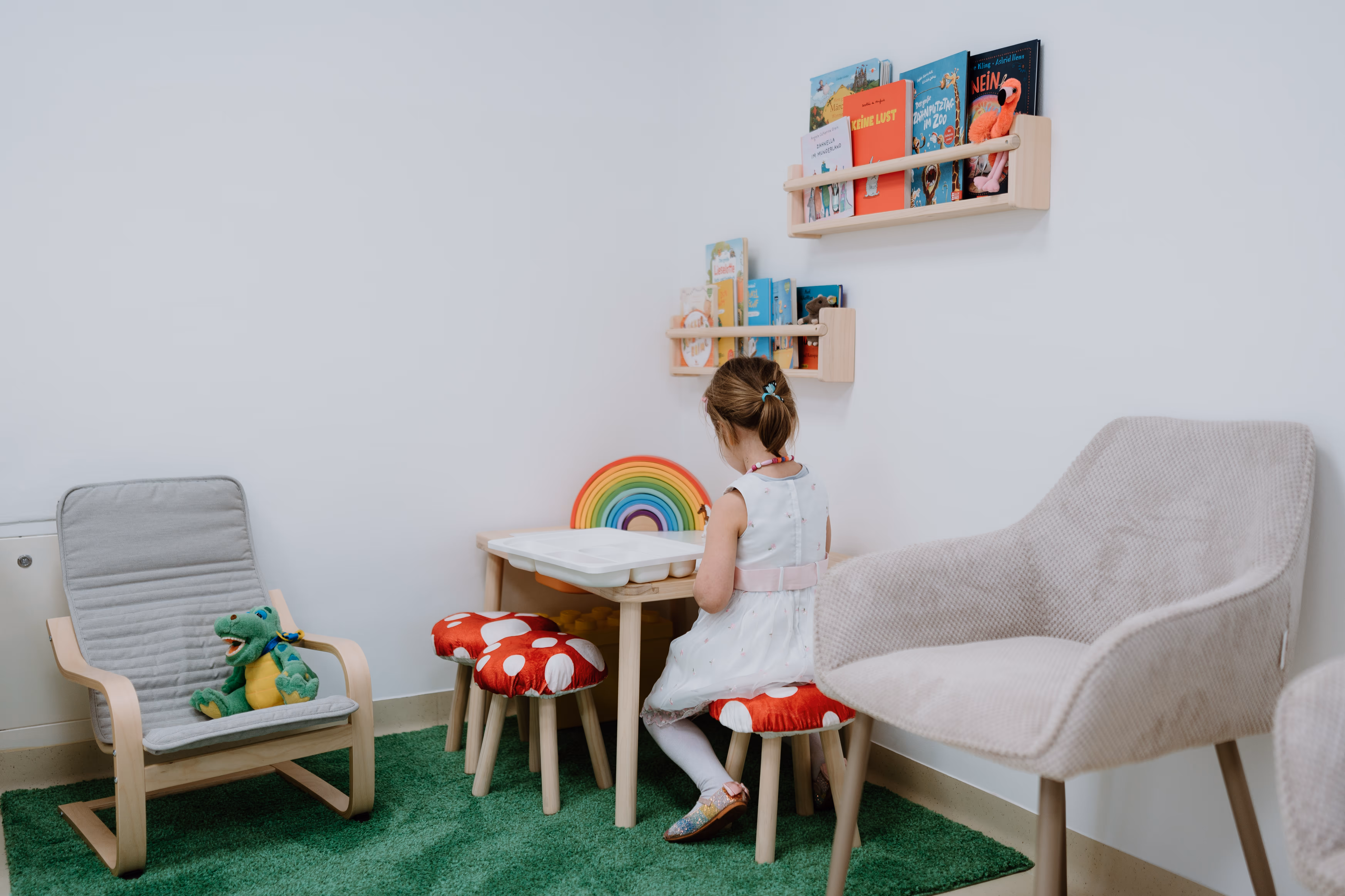 Ein kleines Mädchen in weißem Kleid sitzt auf einem roten Hocker mit weißen Punkten an einem Kinderleichtertisch in einem Raum mit Wandregalen voller Kinderbücher.