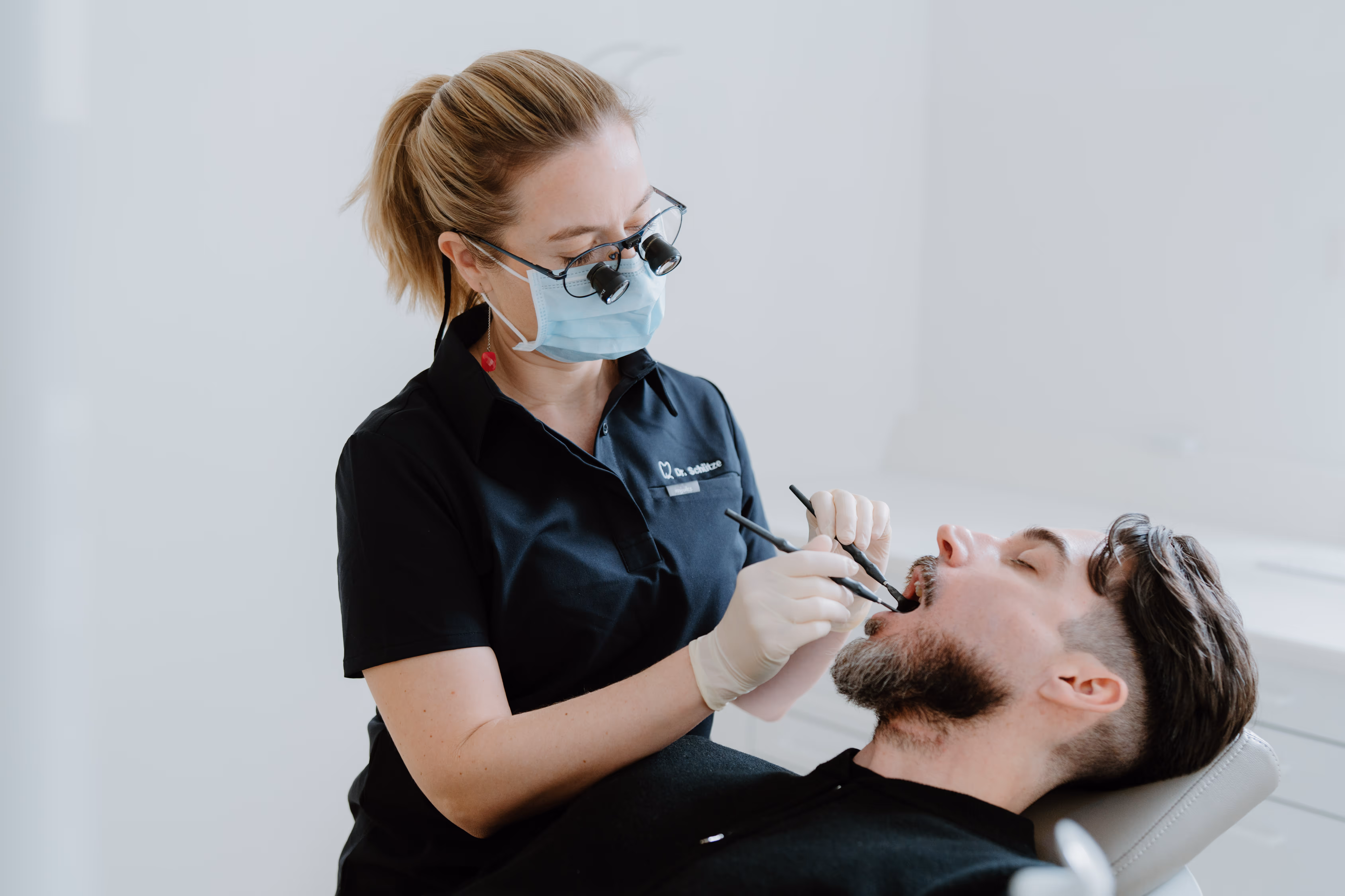 Zahnärztin mit Schutzmaske und Lupenbrille behandelt einen Patienten, der in einem Zahnarztstuhl liegt.
