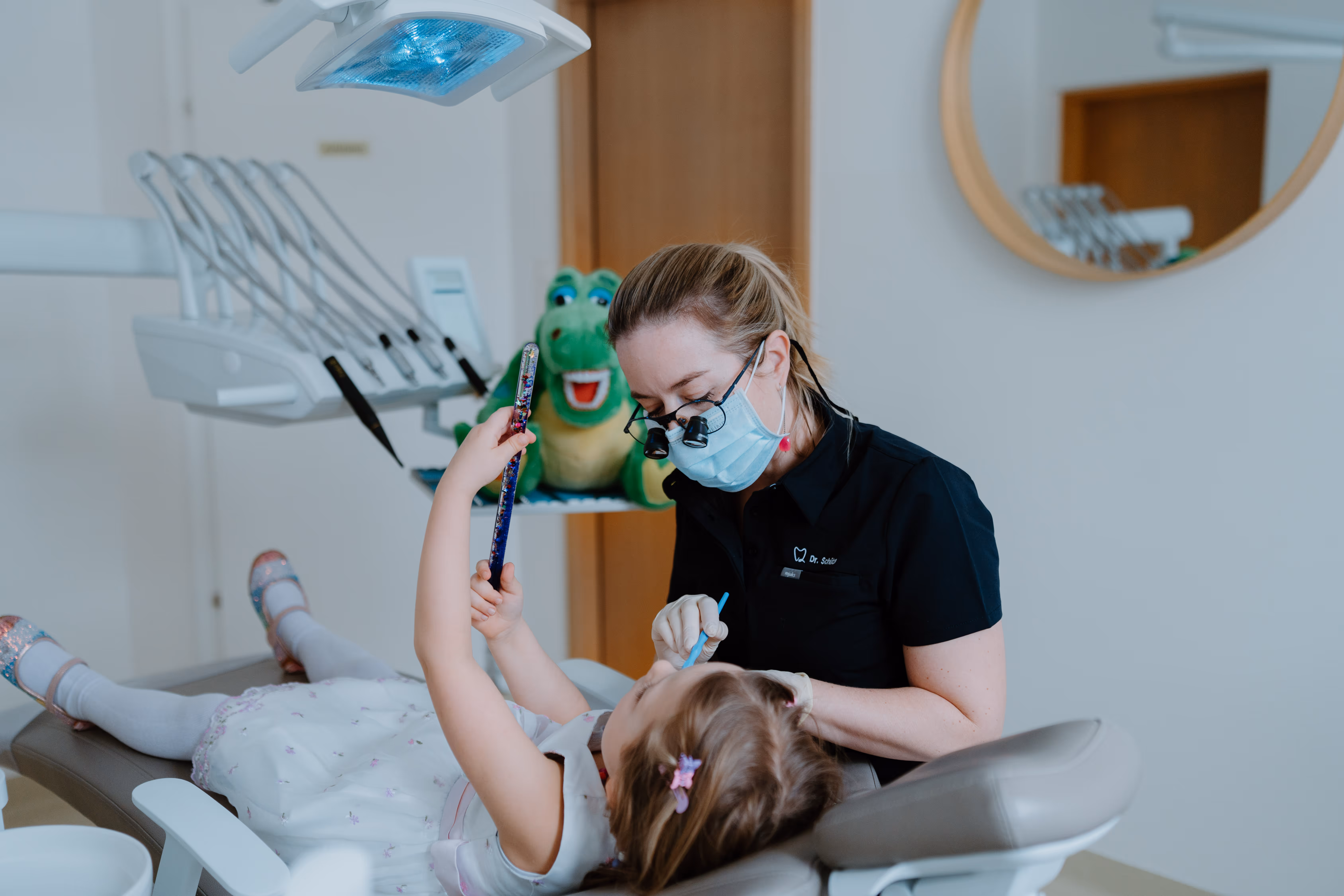 Zahnärztin mit Gesichtsmaske behandelt ein kleines Mädchen in einer Zahnarztpraxis, das Mädchen hält einen magischen Zauberstab.