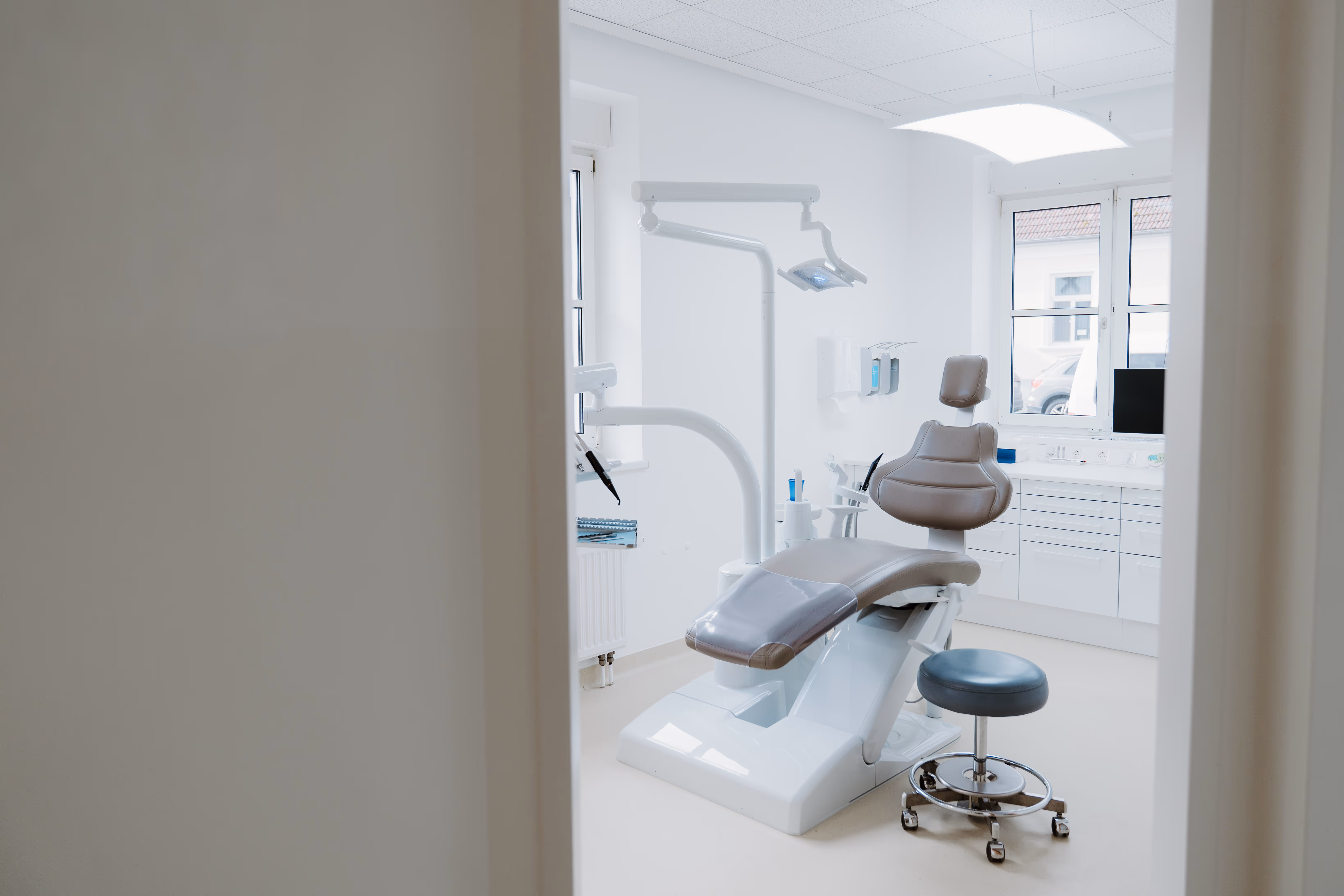 Moderne Zahnarztpraxis mit Zahnarztstuhl, Beistellhocker und großen Fenstern.