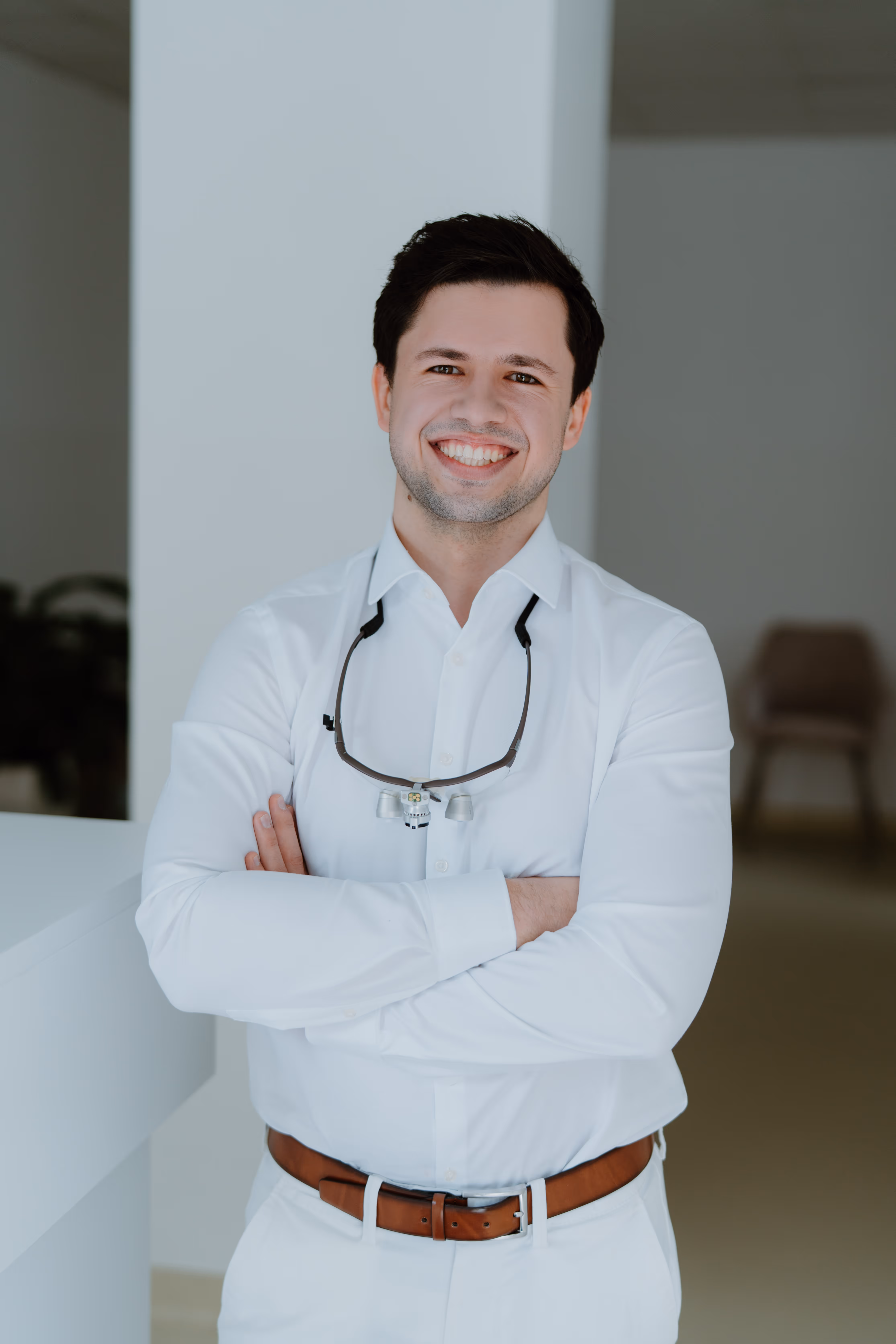 Junger Zahnarzt mit weißem Hemd und braunem Gürtel lächelt, trägt eine zahnärztliche Lupe um den Hals.