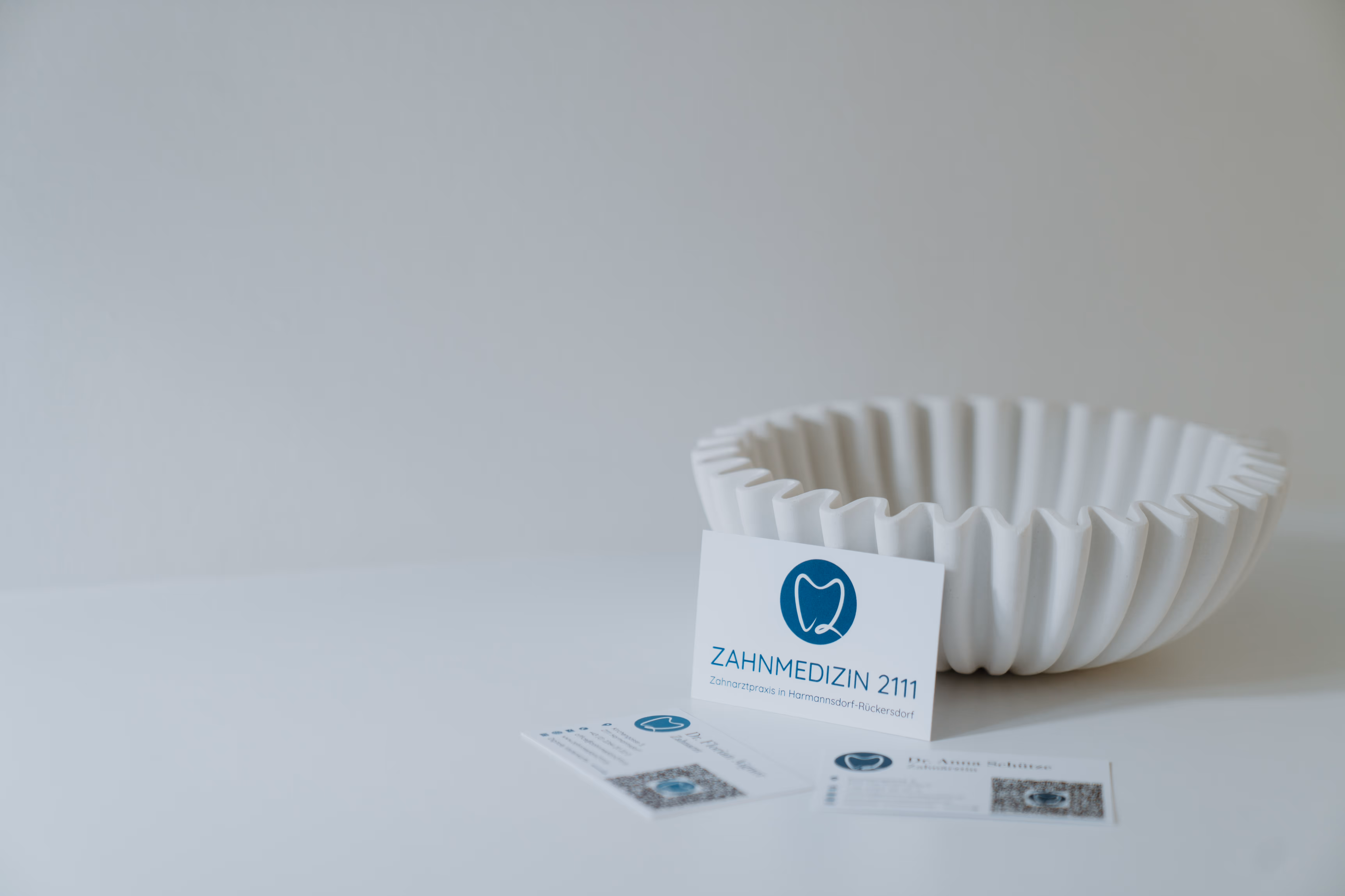 Visitenkarten von Zahnmedizin 2111 mit Zahnlogo vor einer weißen, wellenförmigen Schale auf weißem Hintergrund.