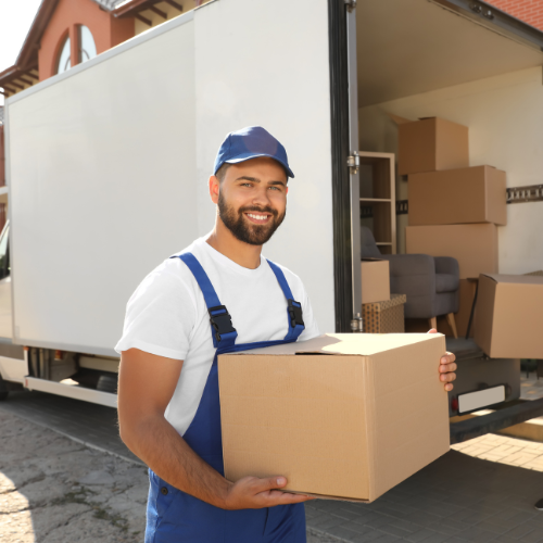 Smiling mover in blue cap and overalls holding a cardboard box in front of a moving truck with packed boxes inside.