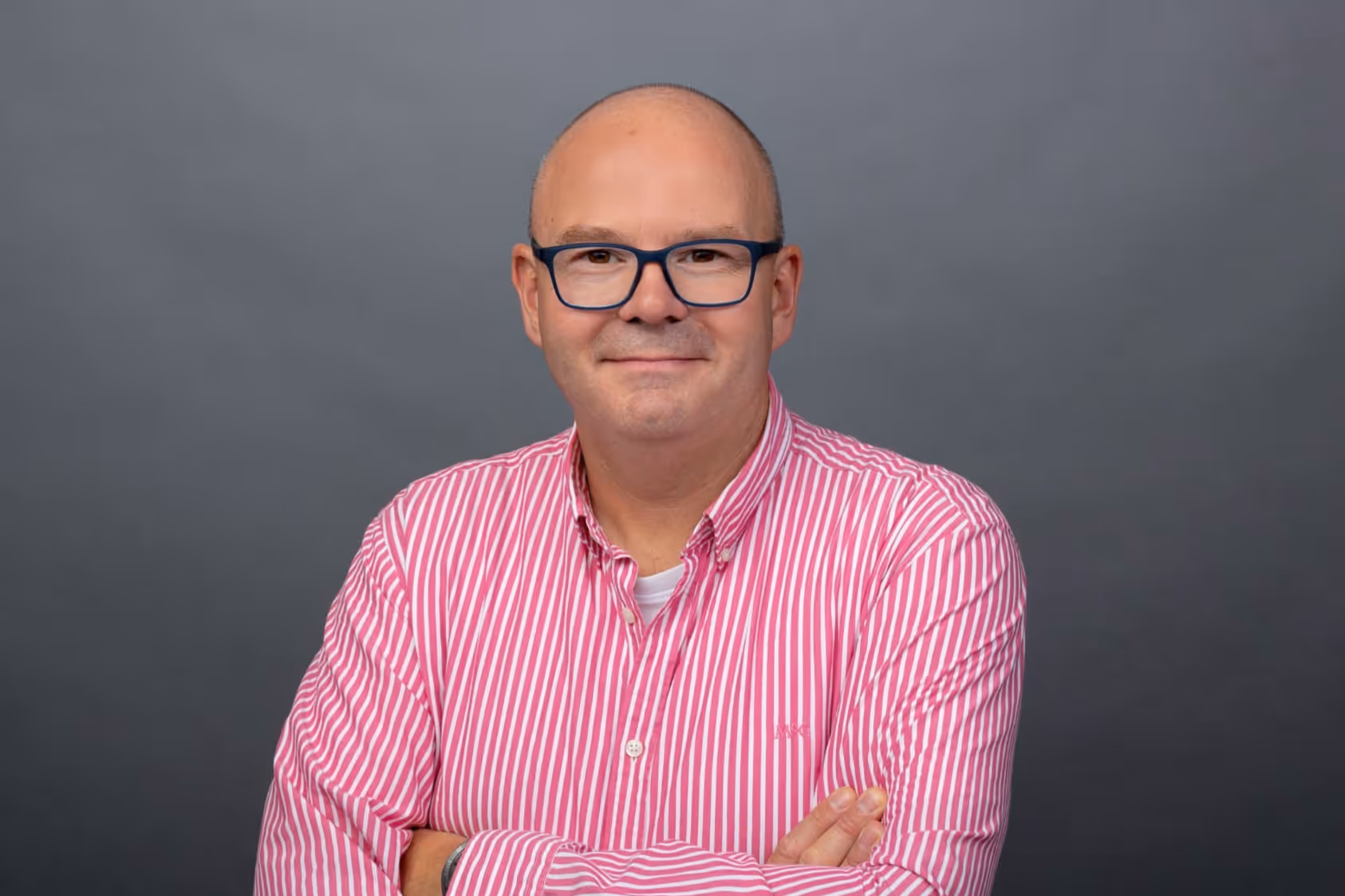 Man met een roze gestreept overhemd en bril, armen gekruist, voor een grijze achtergrond.