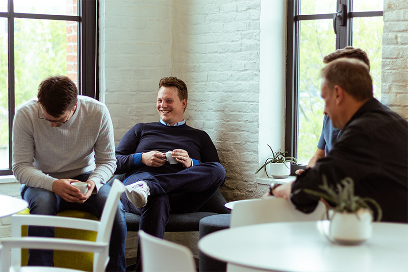 Four men sitting and chatting in a bright room with large windows, holding coffee cups and smiling.
