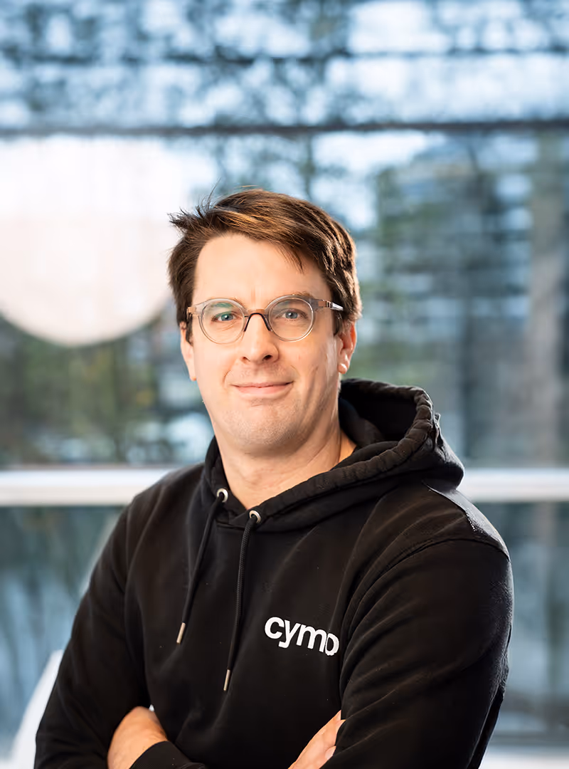 Man wearing glasses and a black hoodie with the word 'cymp' smiling with arms crossed indoors.